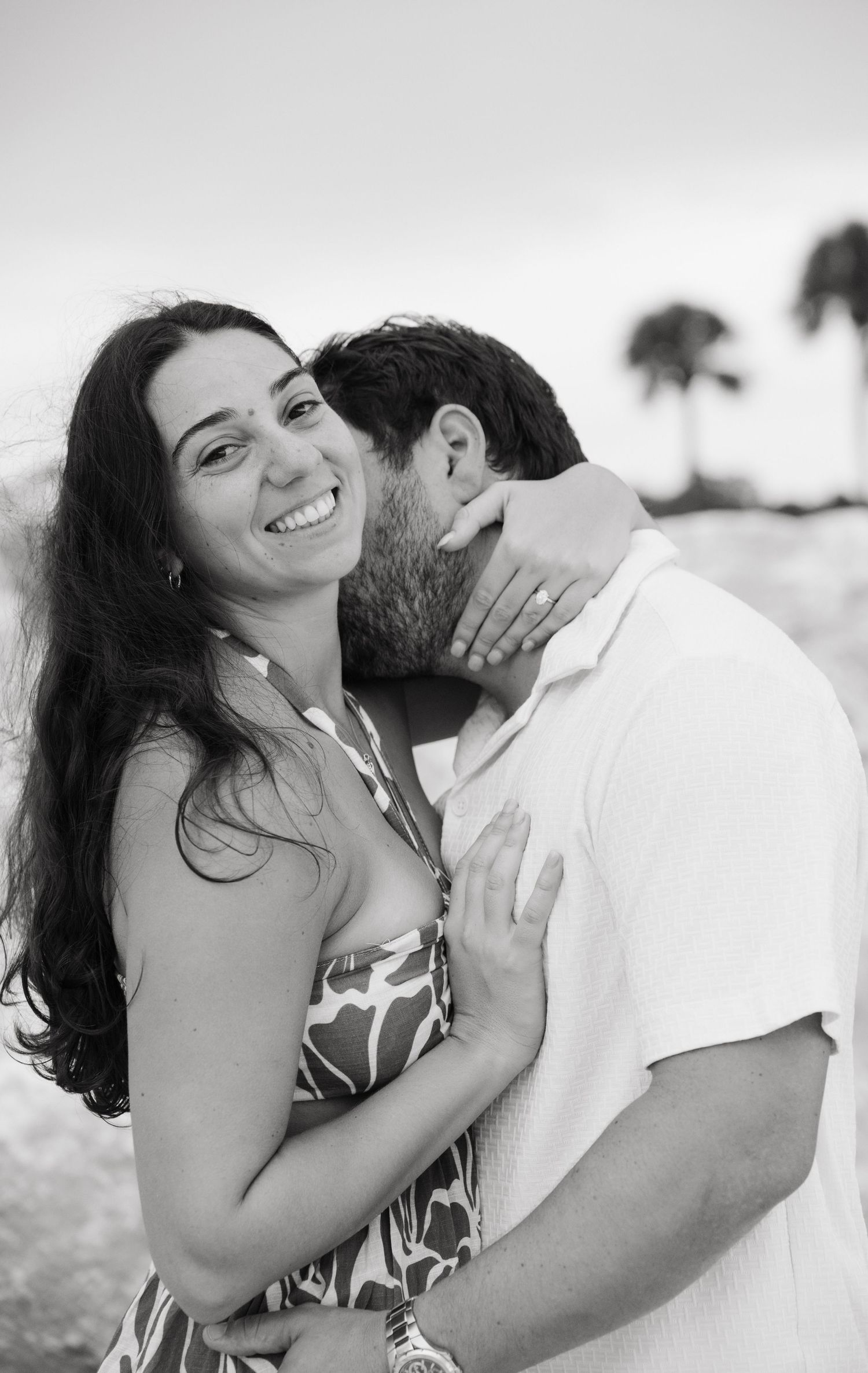 Black and white beach photo of a smiling woman being kissed on the cheek by a man, with palm trees in background.