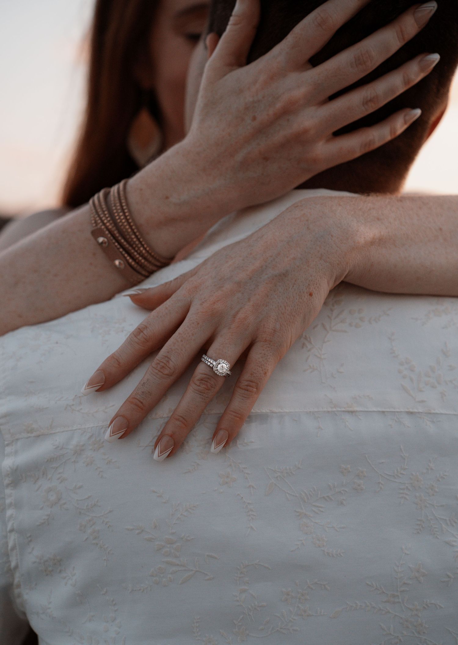 Close-up of a woman's hand with a diamond engagement ring and gold bracelet, resting on a man's white shirt.