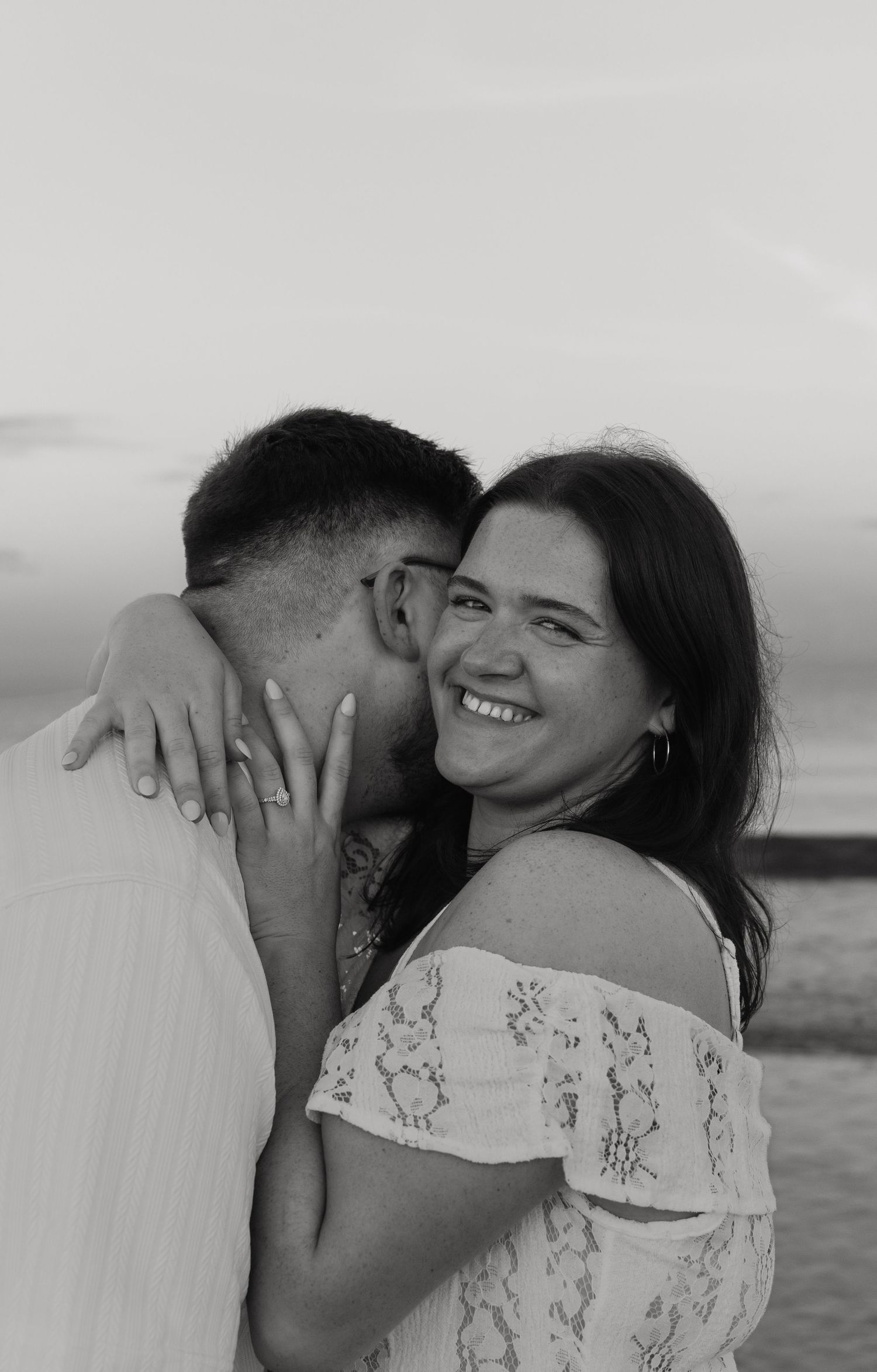 Black and white photo of a smiling woman in lace top being kissed on cheek by man outdoors at sunset.