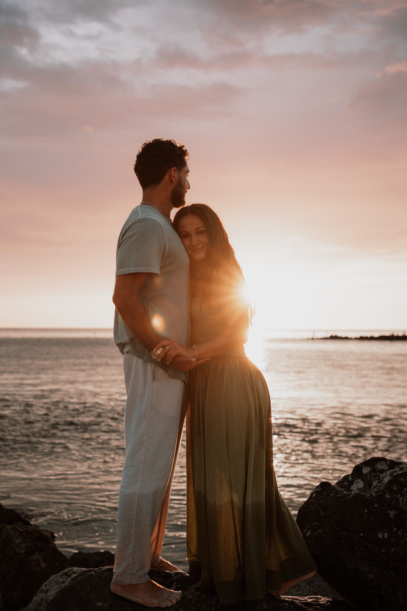Couple embracing at sunset on rocky beach, golden light glowing behind them, woman in green dress, man in white outfit.