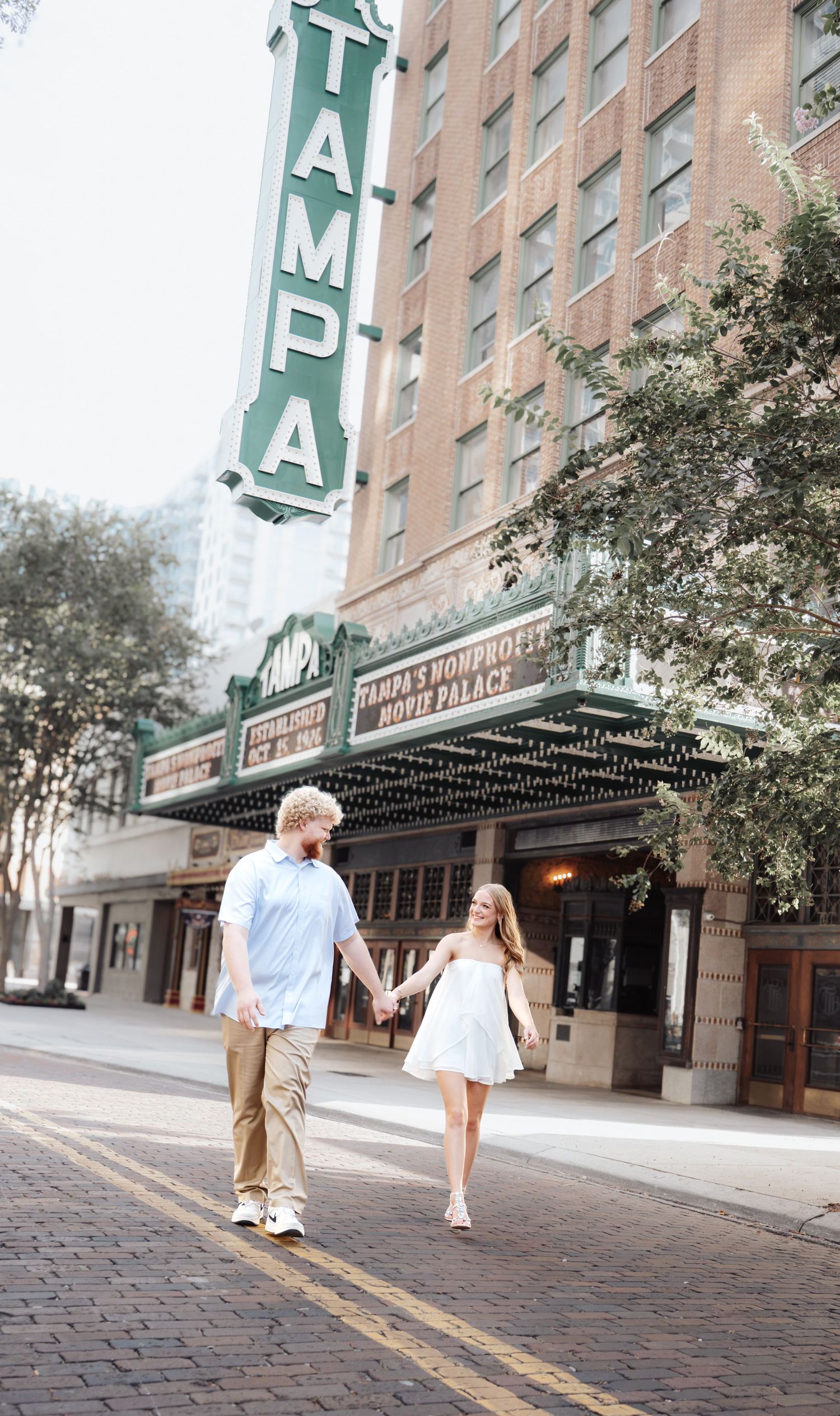 Couple holding hands walking in front of the historic Tampa Theatre marquee on a sunny day.