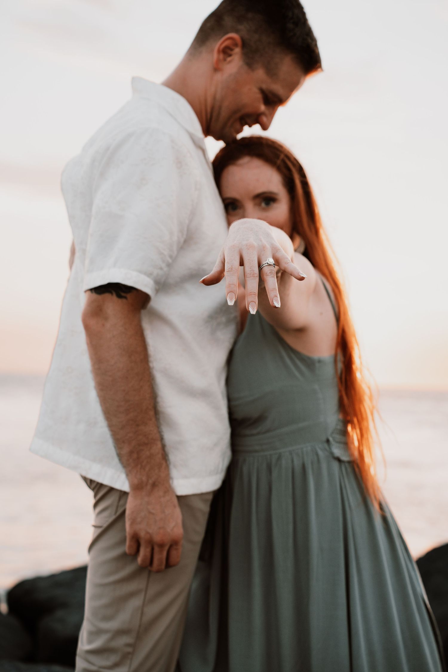 Couple embracing at sunset, woman in sage green dress showing off her diamond engagement ring.