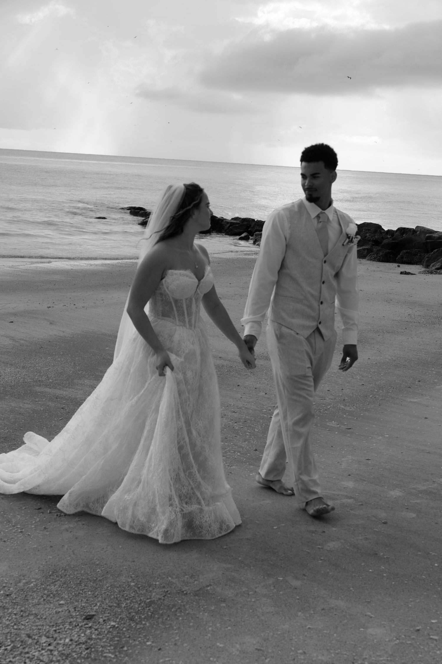 Black and white photo of a bride and groom holding hands walking on a beach in wedding attire.