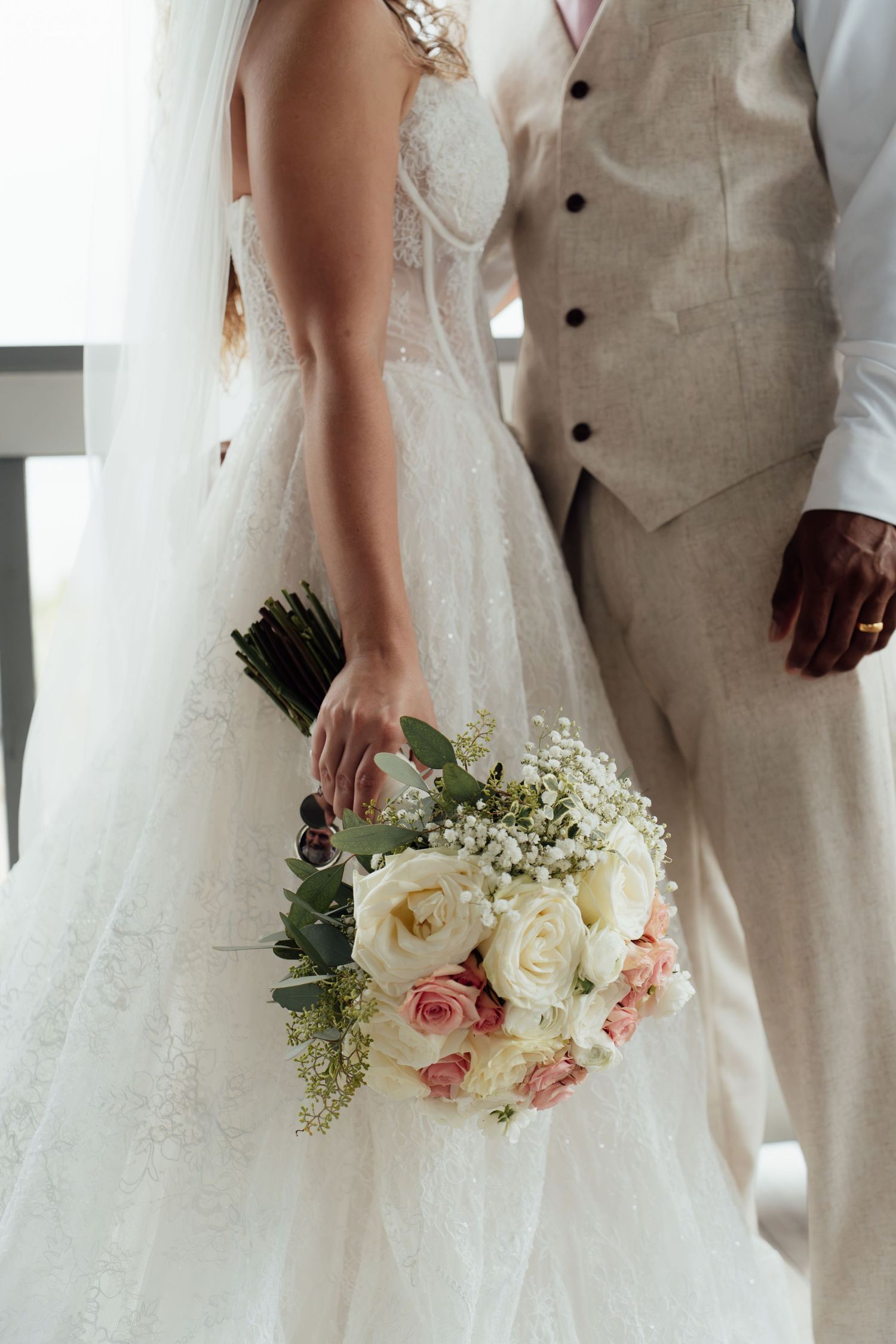 Bride in lace gown holding pink and white rose bouquet beside groom in beige suit at wedding ceremony.