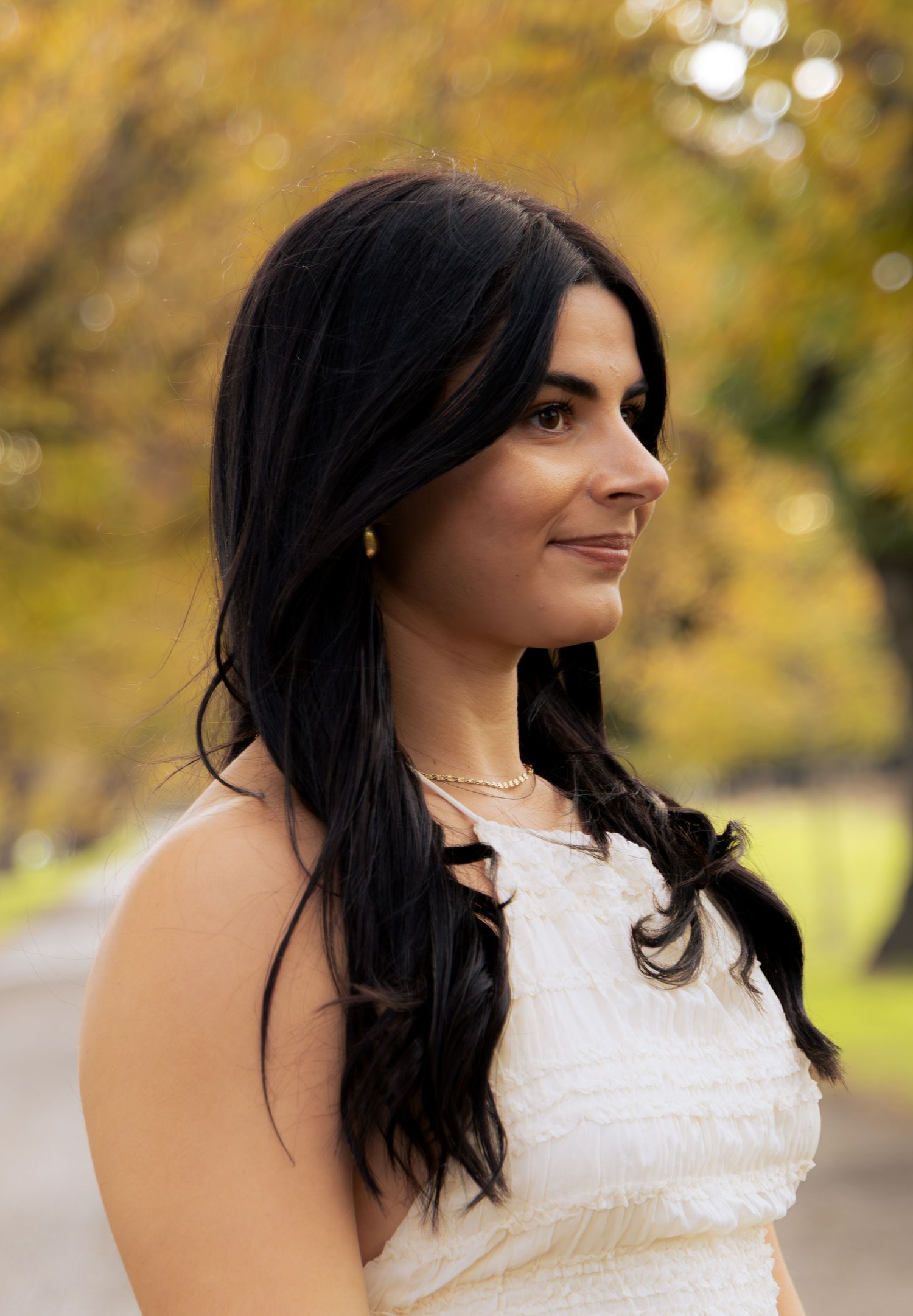 Young woman with long black hair in white dress gazing sideways on a tree-lined autumn path.