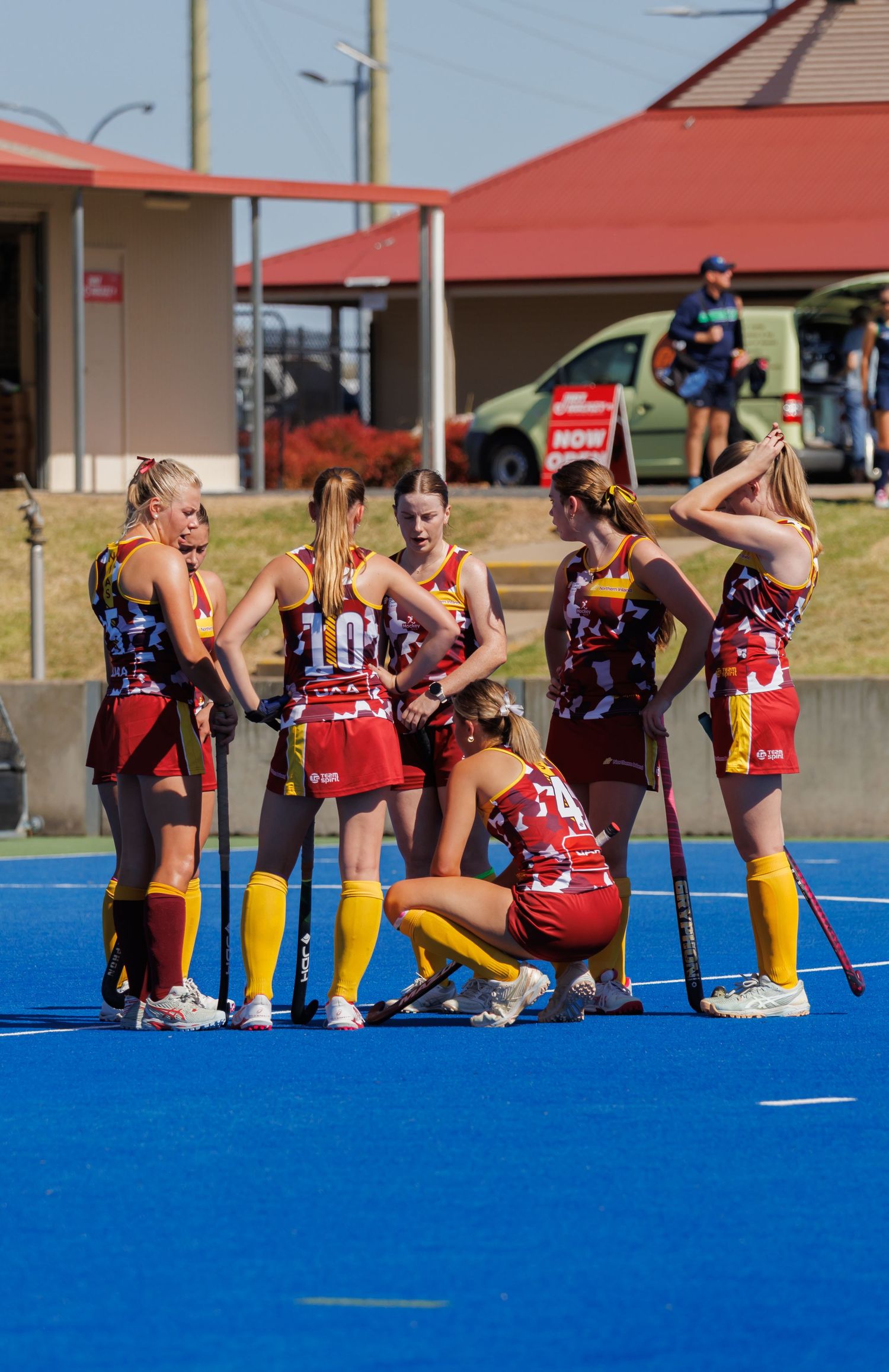 A girls field hockey team huddles together on a blue turf court during a competitive match.