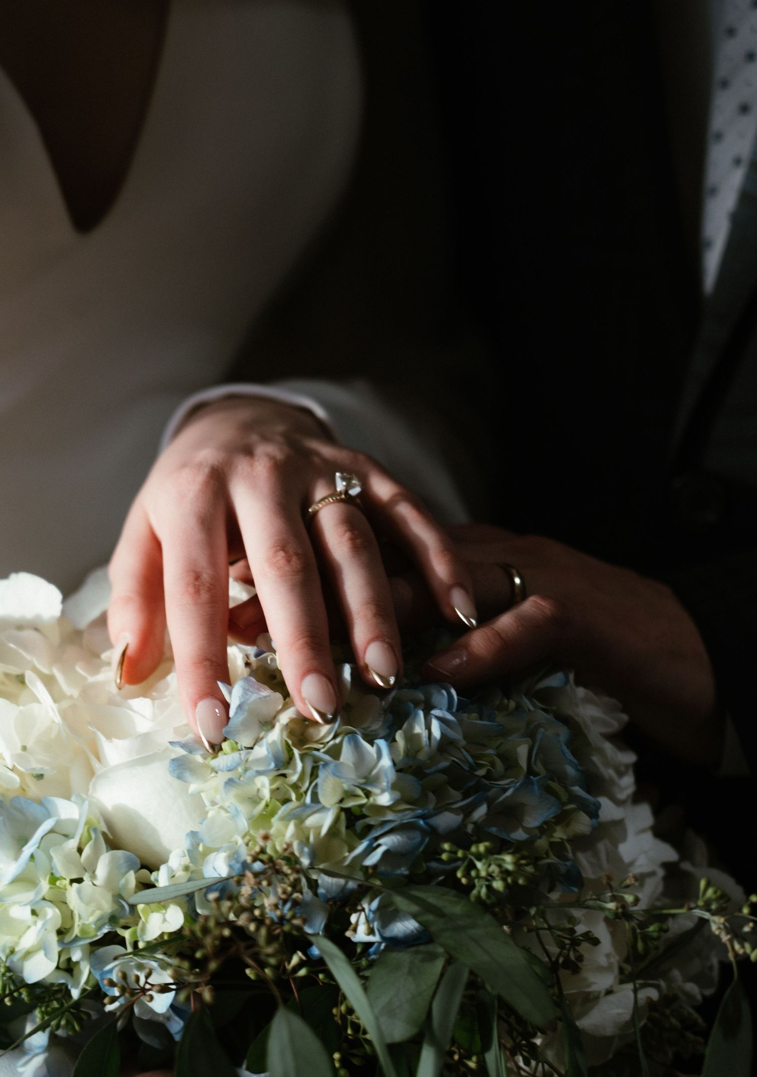 Close-up of newlyweds hands resting on a white and blue hydrangea bridal bouquet with wedding rings visible.