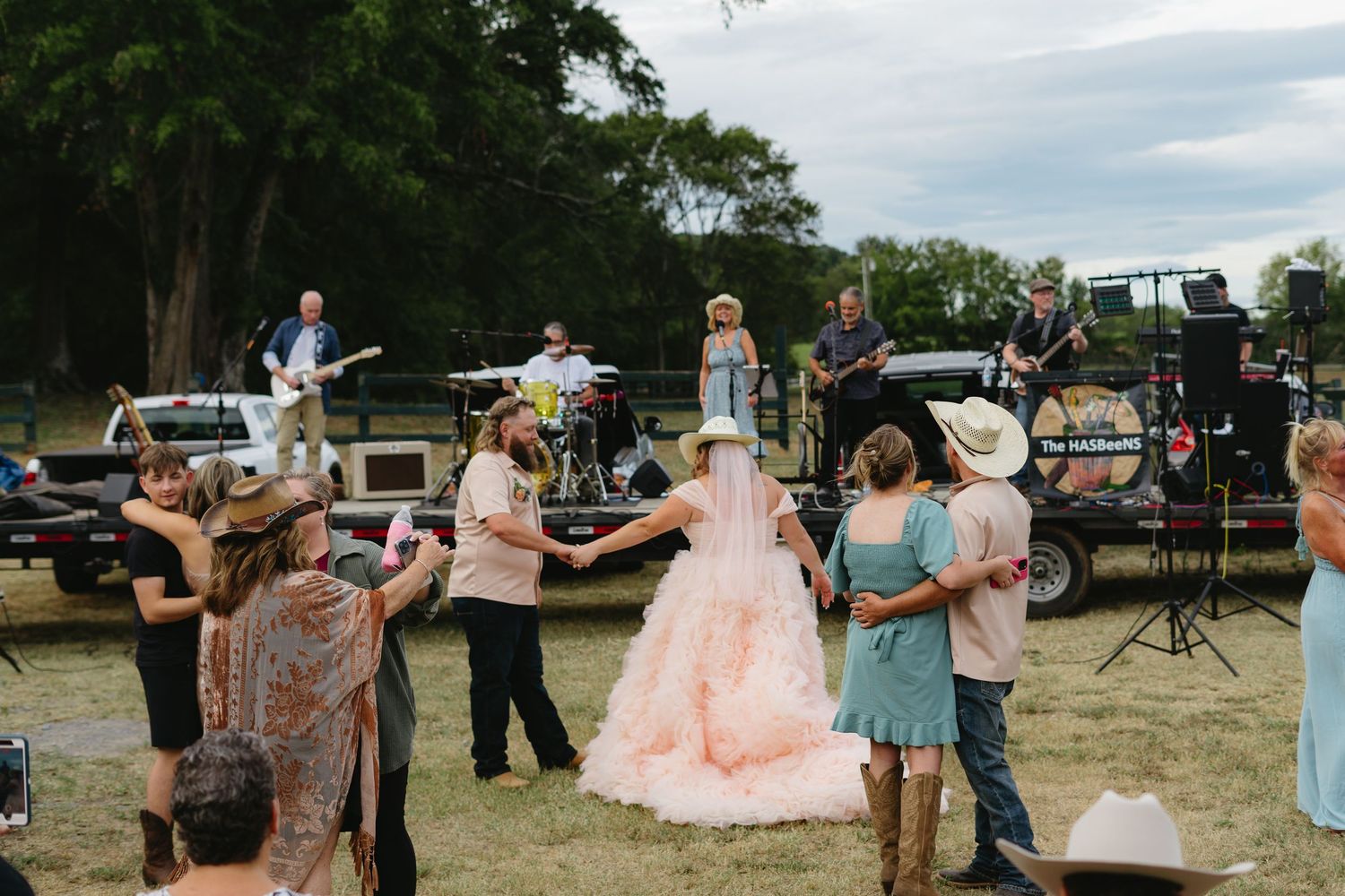 Guests dance outdoors at a country wedding reception with a live band performing on a rustic stage.