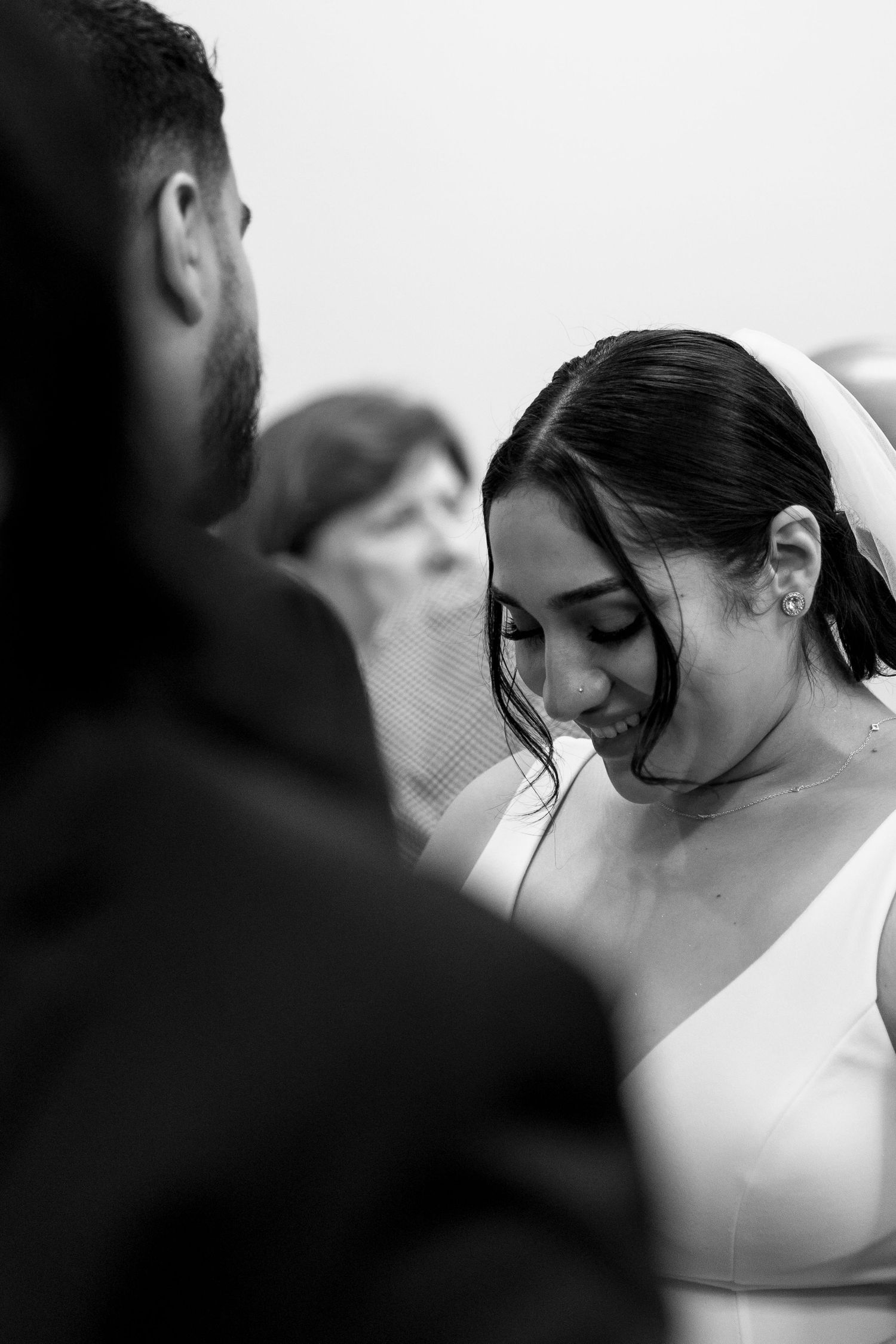 A smiling bride in a white gown and veil looks down during her wedding ceremony in black and white.