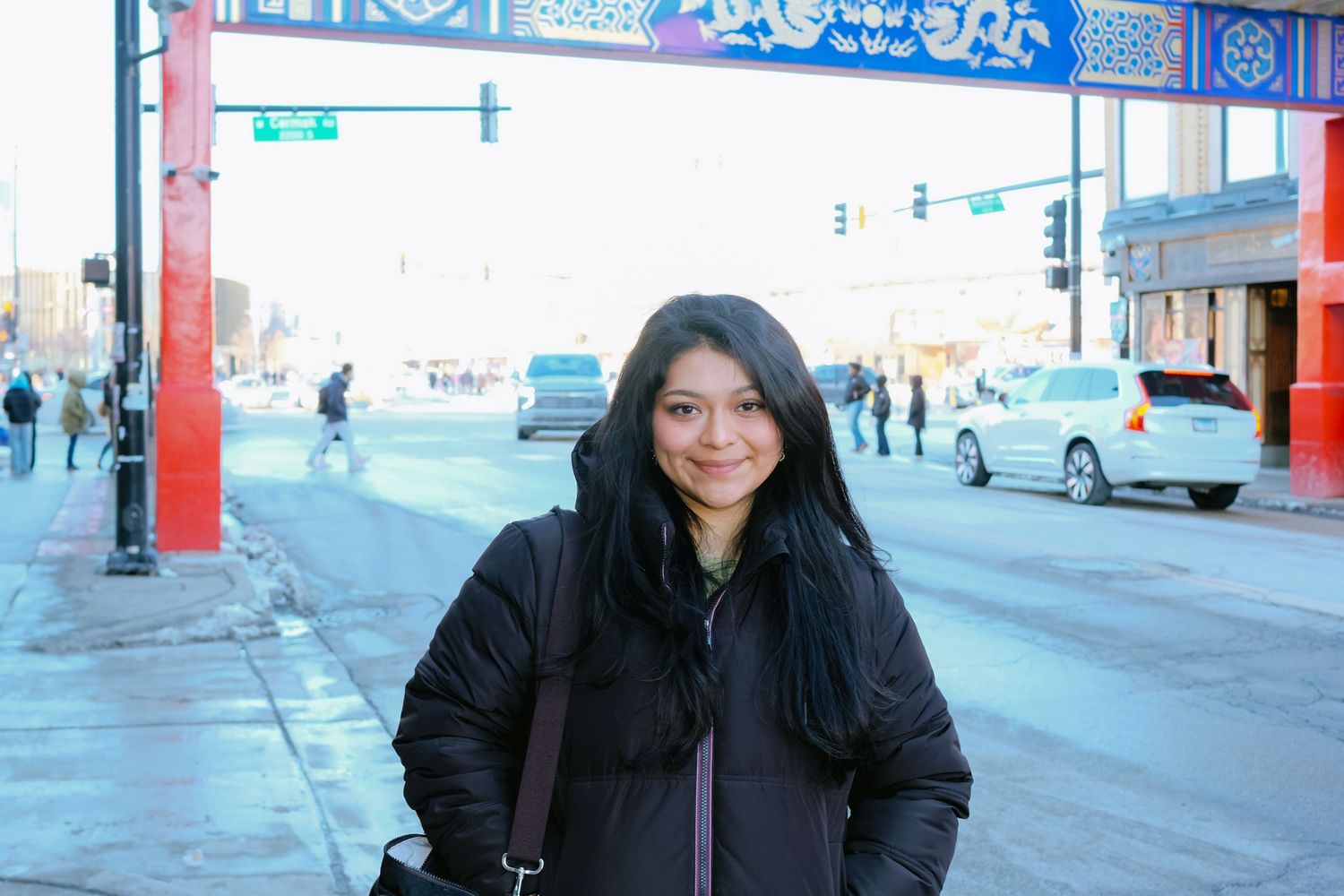 A smiling woman in a black puffer jacket stands on a busy urban street corner in what appears to be Chinatown.