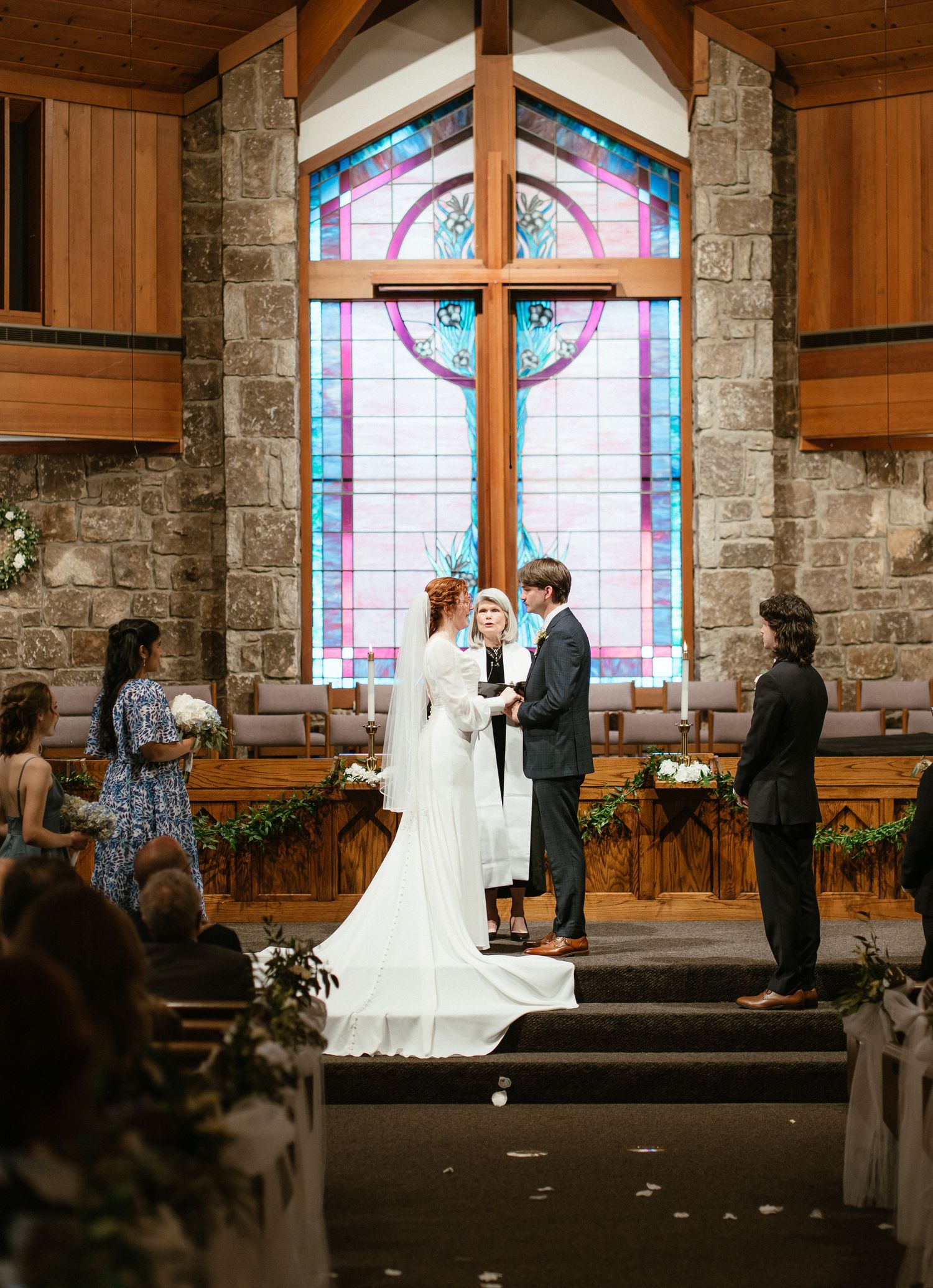 Bride and groom exchange vows at a church altar beneath a stunning stained glass window.