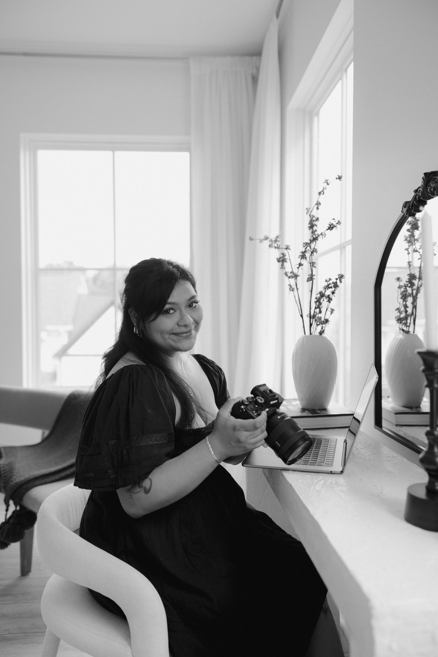 Black and white portrait of woman holding camera at desk by window.