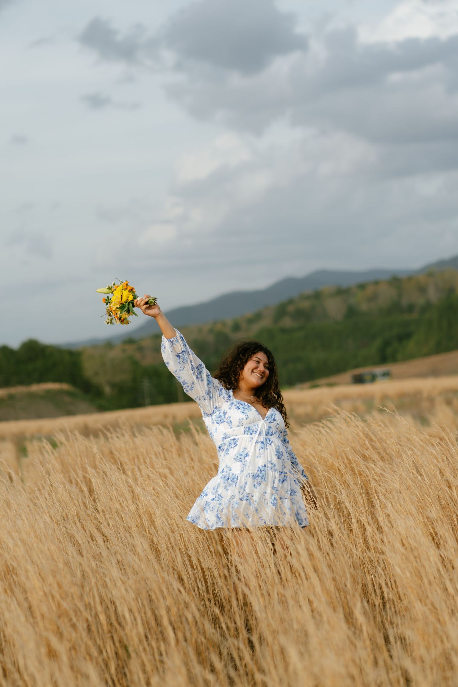 Young woman in blue floral white dress raises yellow sunflower bouquet in golden wheat field with mountains behind.