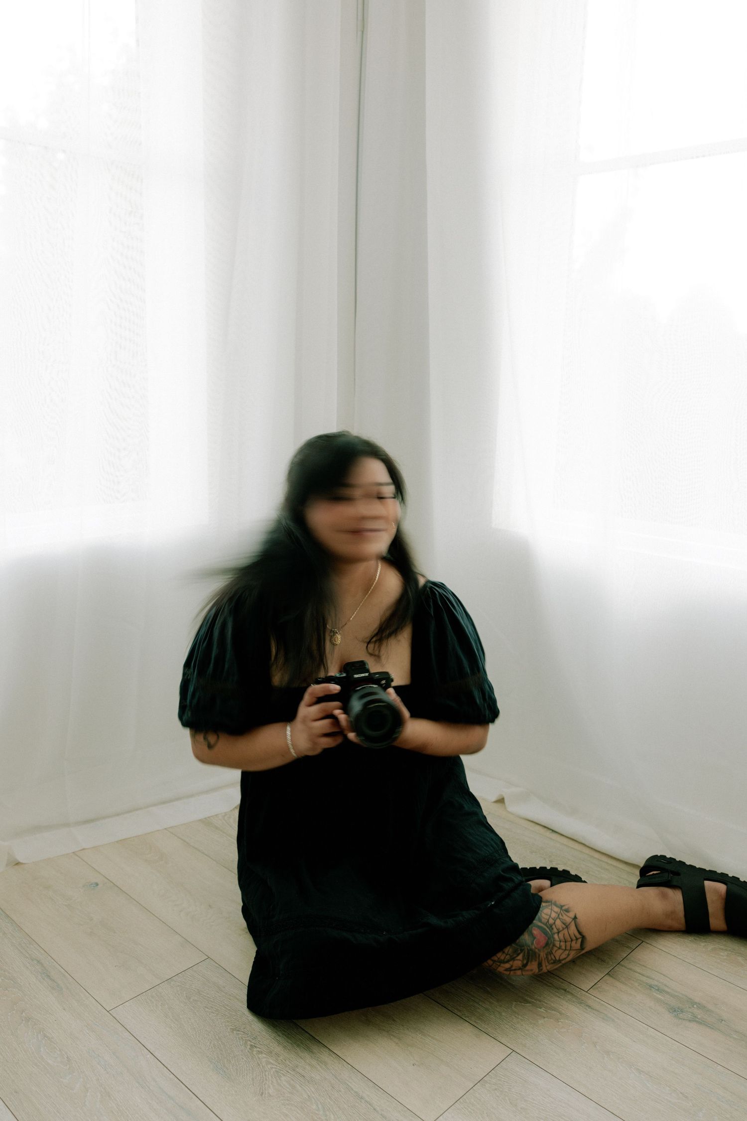 Woman in black dress sitting on floor holding camera near white curtains.