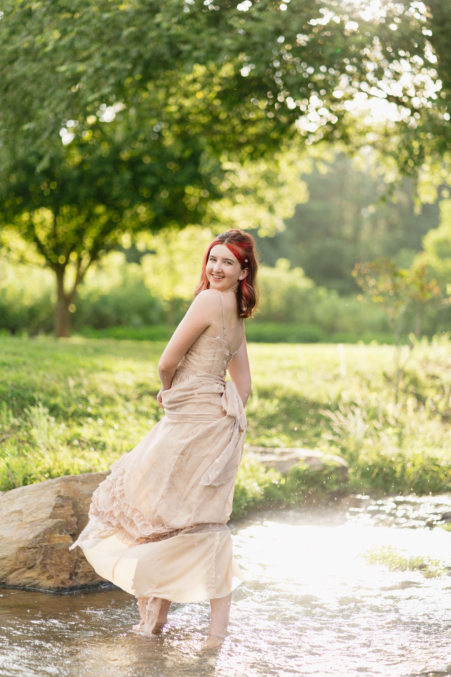 A smiling redhead woman in a flowing beige gown stands in a sunlit creek surrounded by green trees.