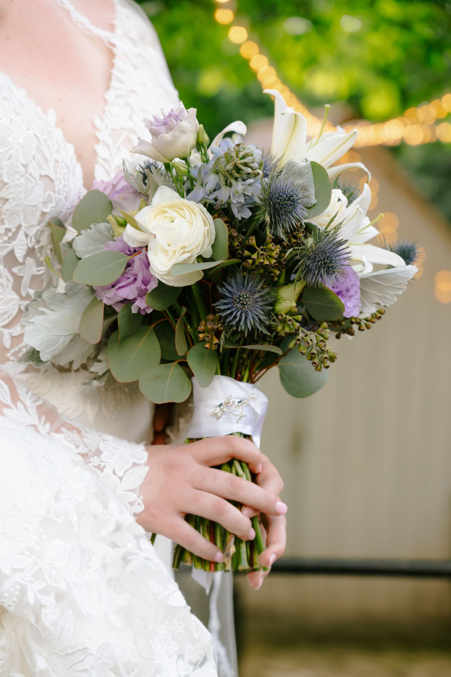 Bride holds a lush bouquet of white roses, purple flowers, and thistles wrapped in white ribbon.