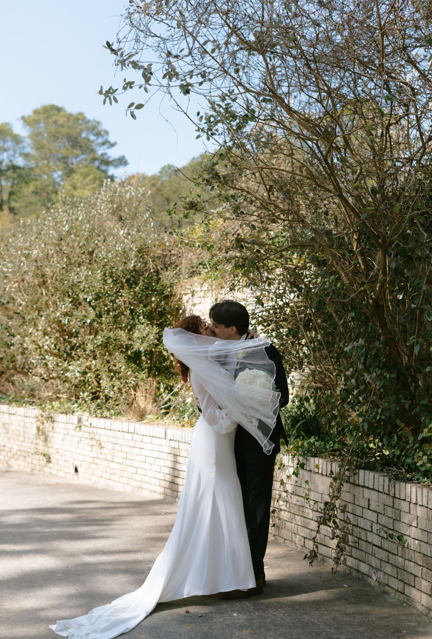 A bride and groom share a romantic kiss outdoors surrounded by lush green foliage on their wedding day.