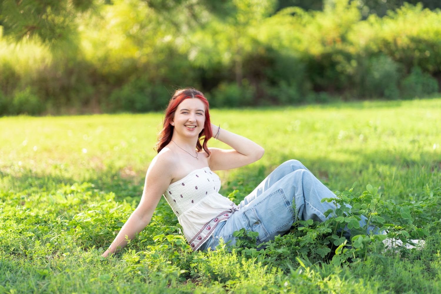 A smiling redhead girl in a white tube top and jeans sits relaxed on a lush green lawn.