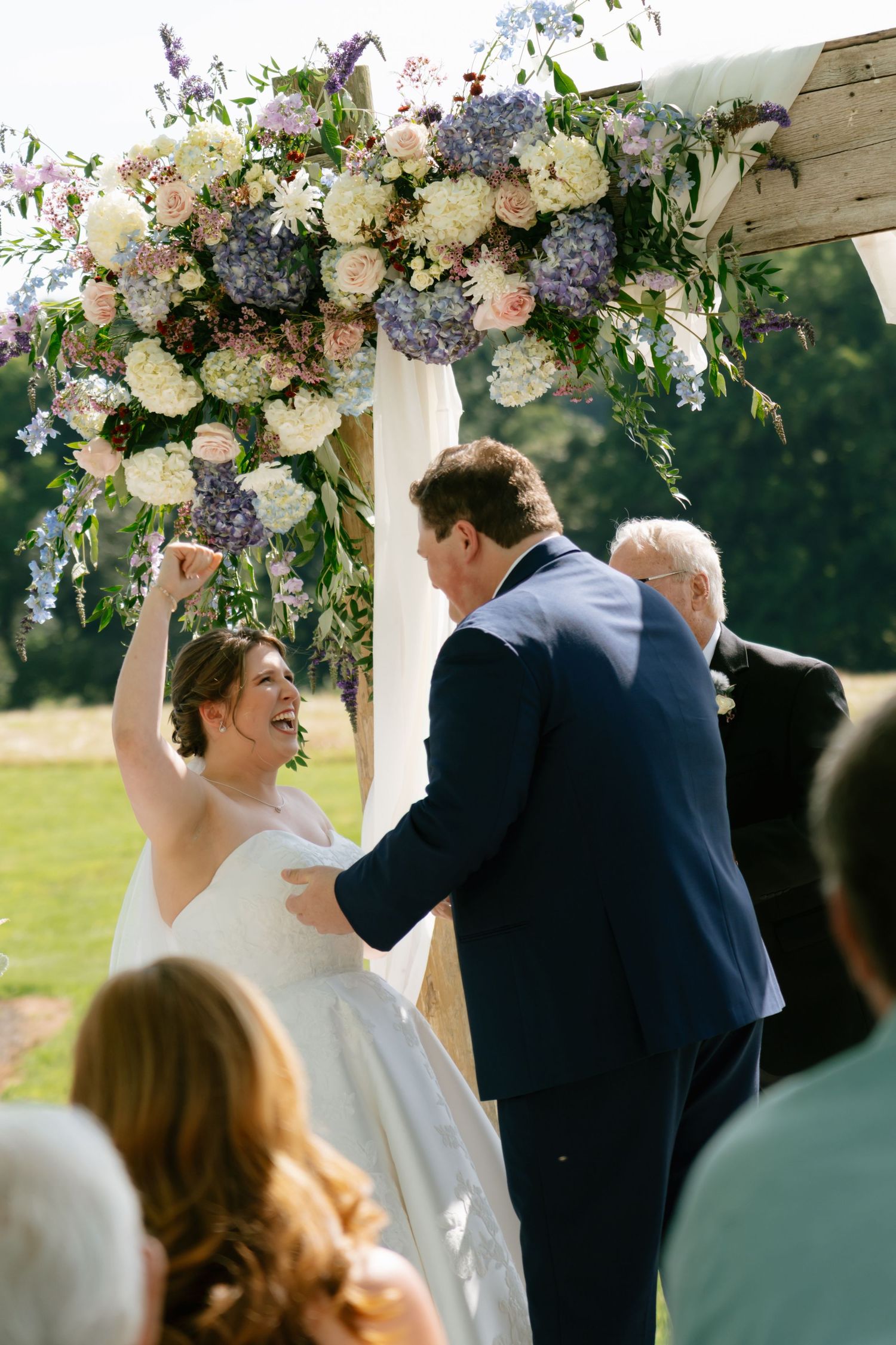 Joyful bride pumps fist at outdoor wedding ceremony under floral arch with purple and white blooms.