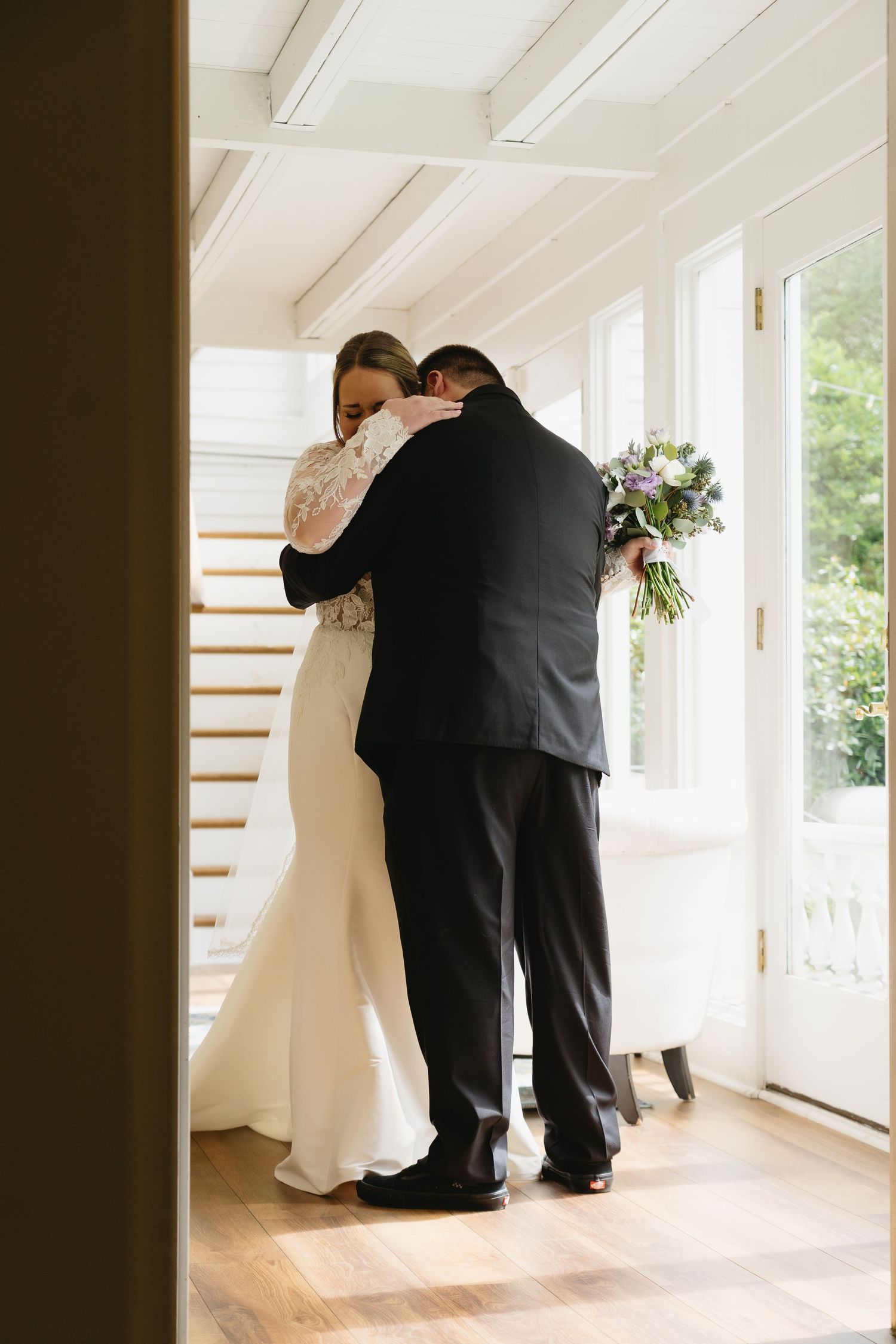 Bride and father share an emotional first look hug near bright windows before the wedding ceremony.