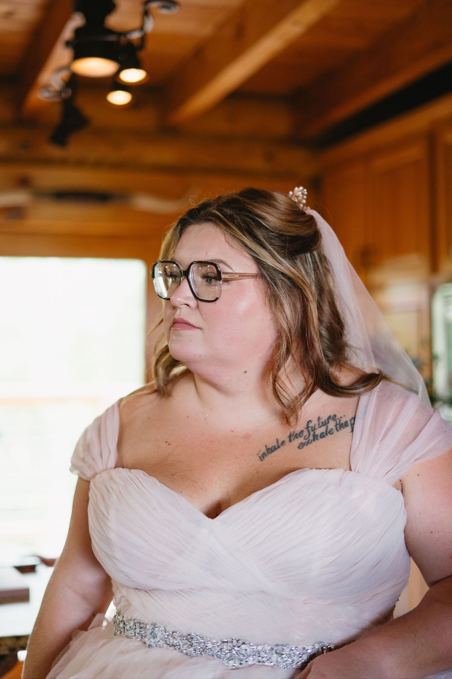 Bride in white gown with jeweled belt, glasses, and veil poses in a rustic wooden venue.