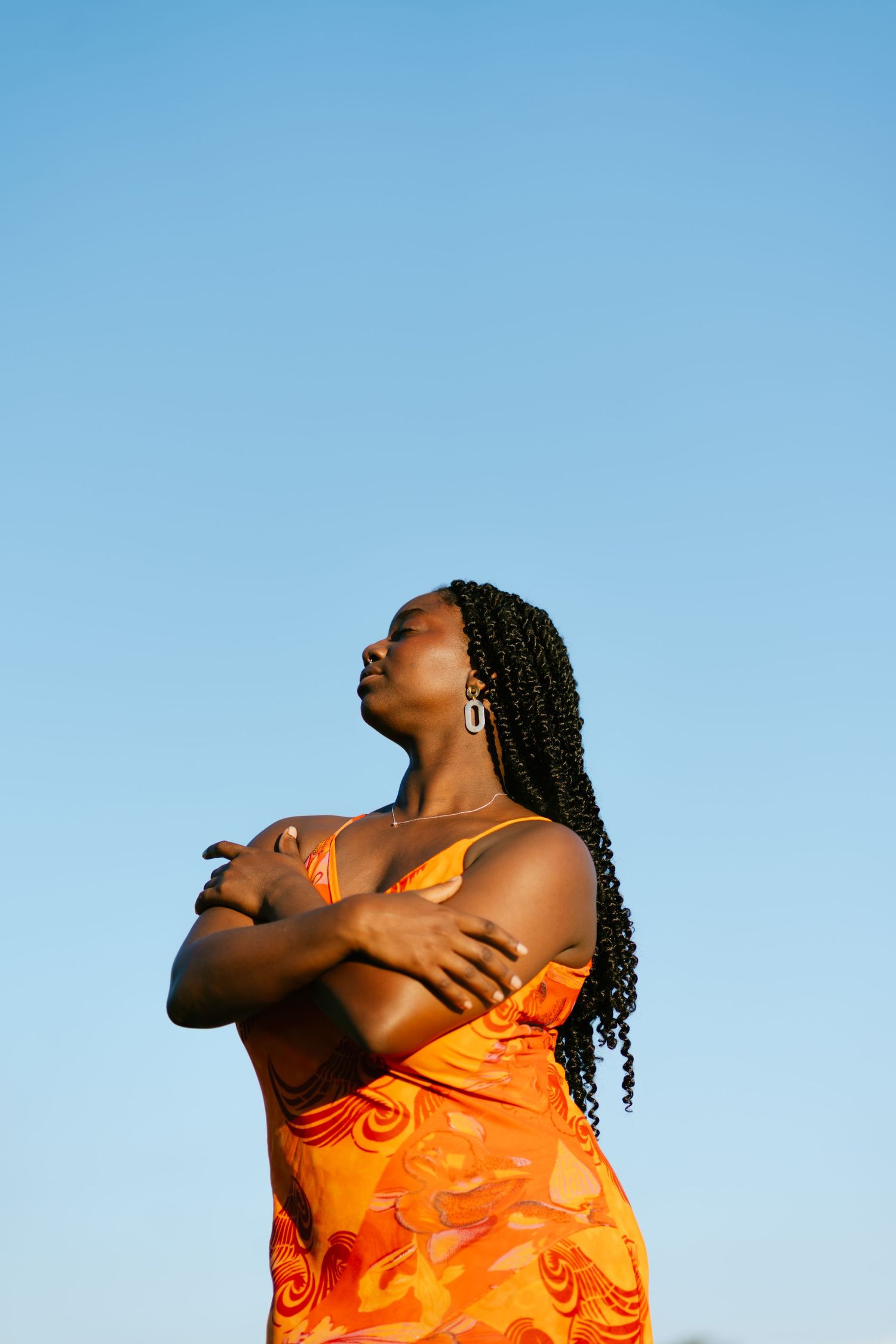 Confident Black woman in orange dress with braids stands arms crossed against clear blue sky.