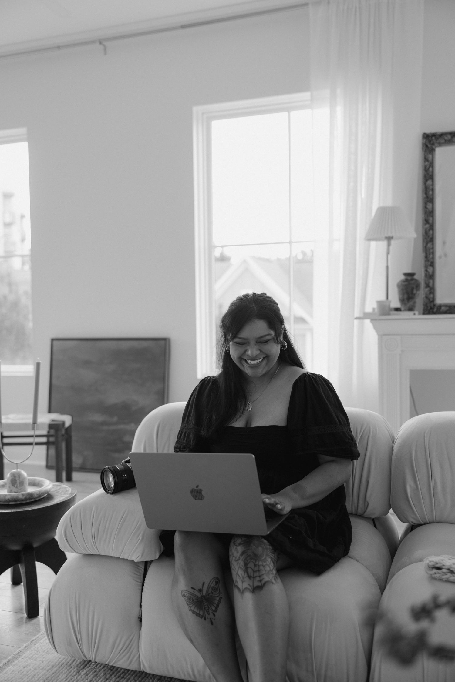 A smiling woman sits on a white armchair using a MacBook laptop in a bright, cozy living room.