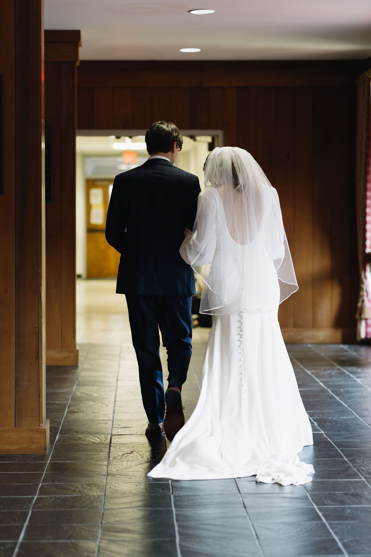 Bride and groom walk hand in hand down a dark hallway, viewed from behind, in wedding attire.