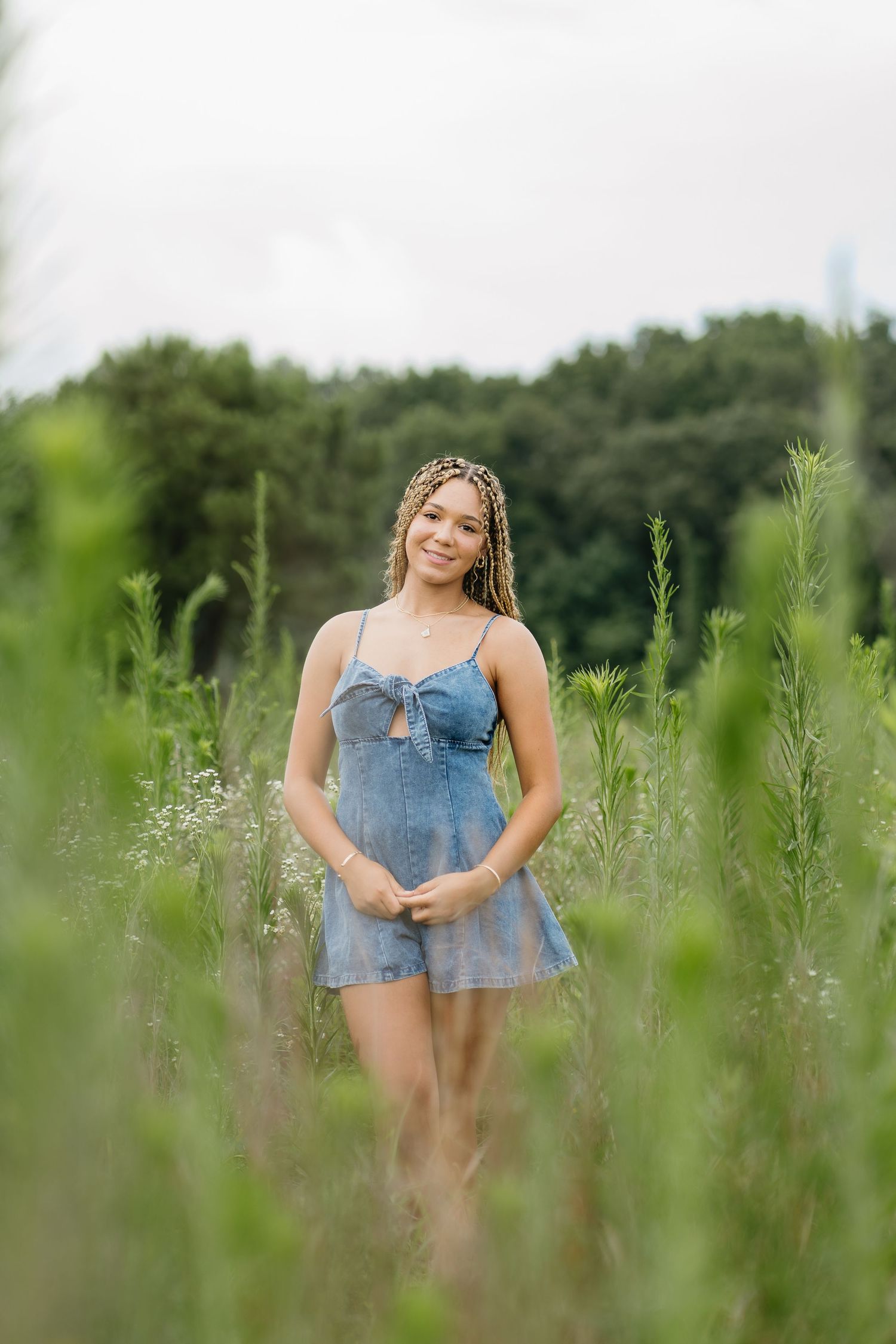 A smiling young woman with braids wears a denim romper while standing in a lush green field.