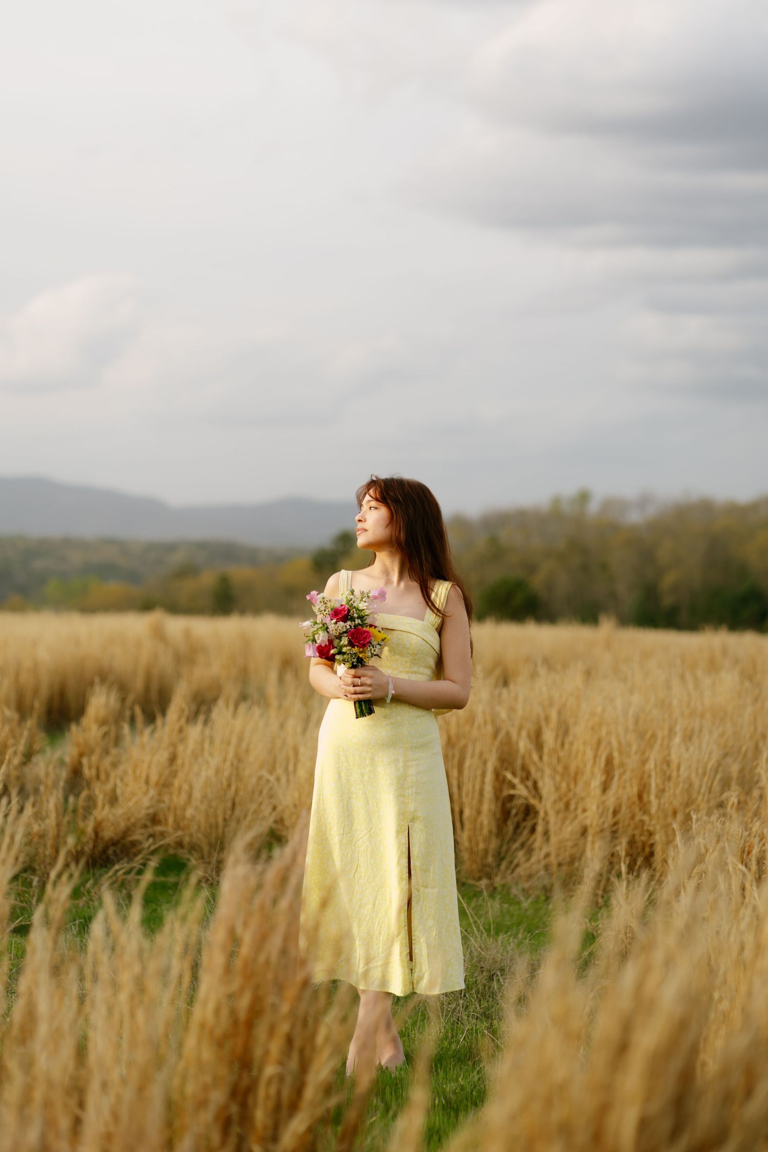 woman in pale yellow midi dress holds pink wildflower bouquet in golden field with mountain backdrop.