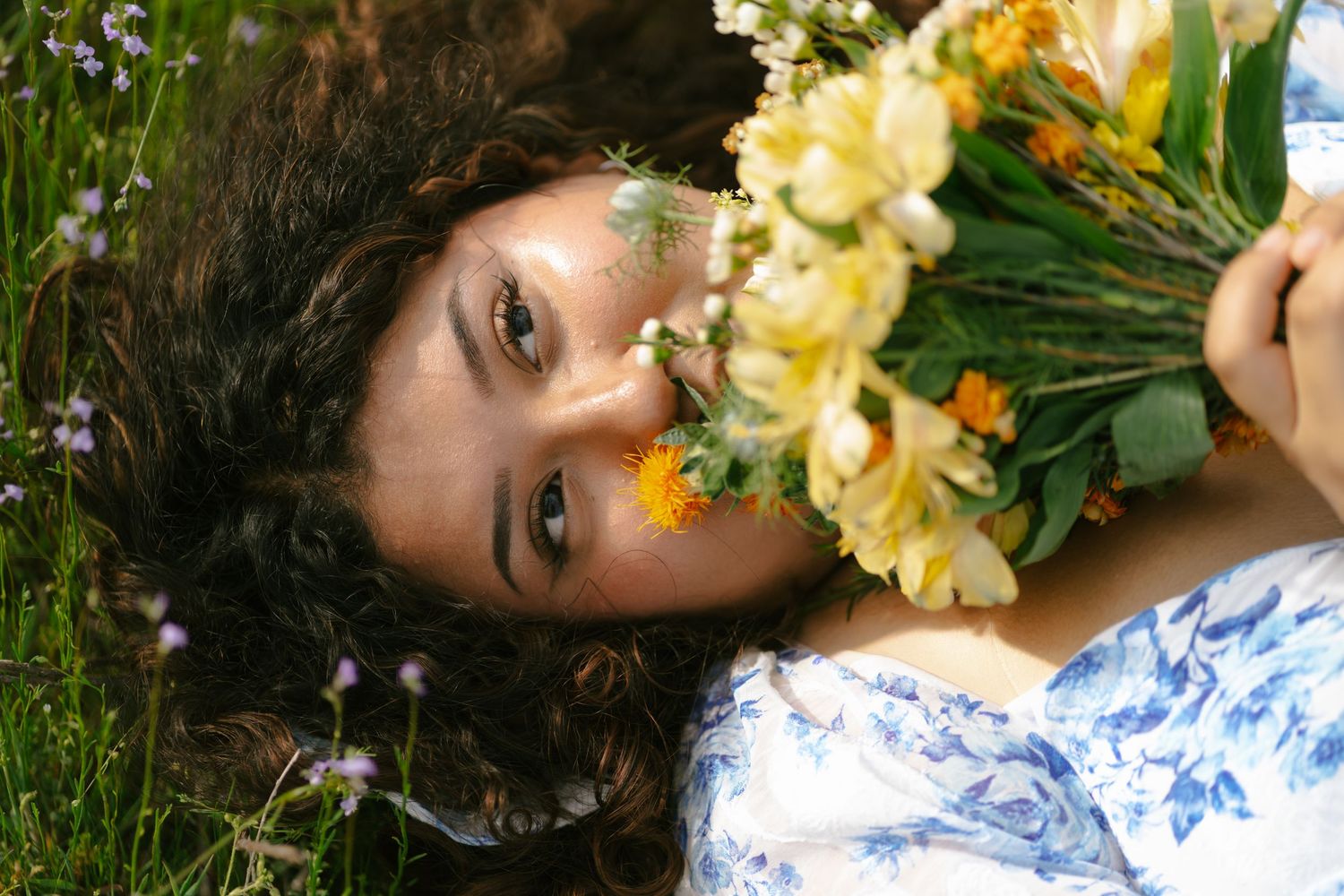 Young woman with curly dark hair lies in flower field holding yellow and white bouquet close to her face.