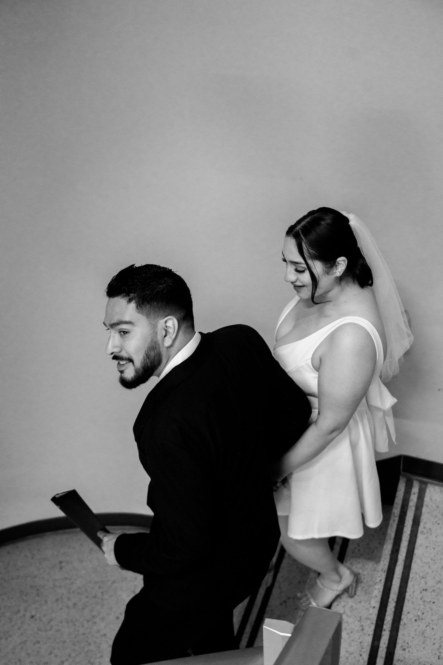 Black and white photo of a bride in white dress and veil standing behind a groom in a suit on stairs.