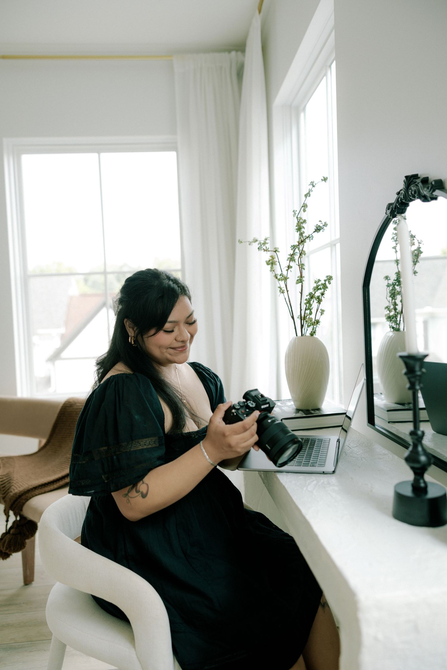 Woman in black dress reviewing photos on camera while sitting at a bright white desk.