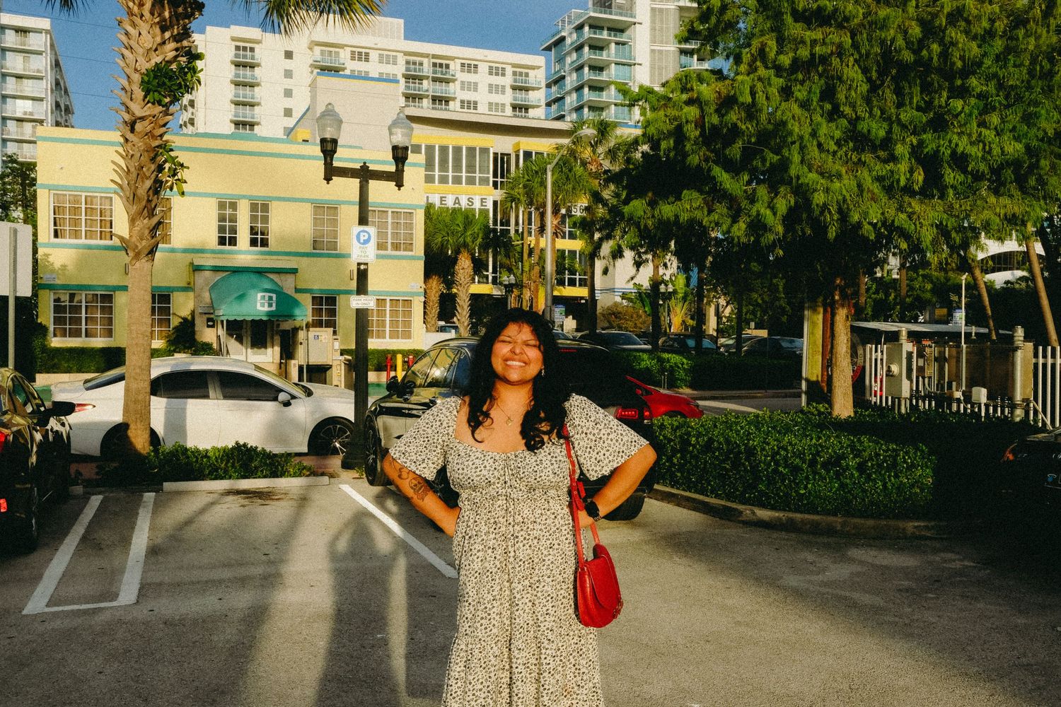 Woman in floral dress smiling outdoors on a sunny street with palm trees and buildings behind her.