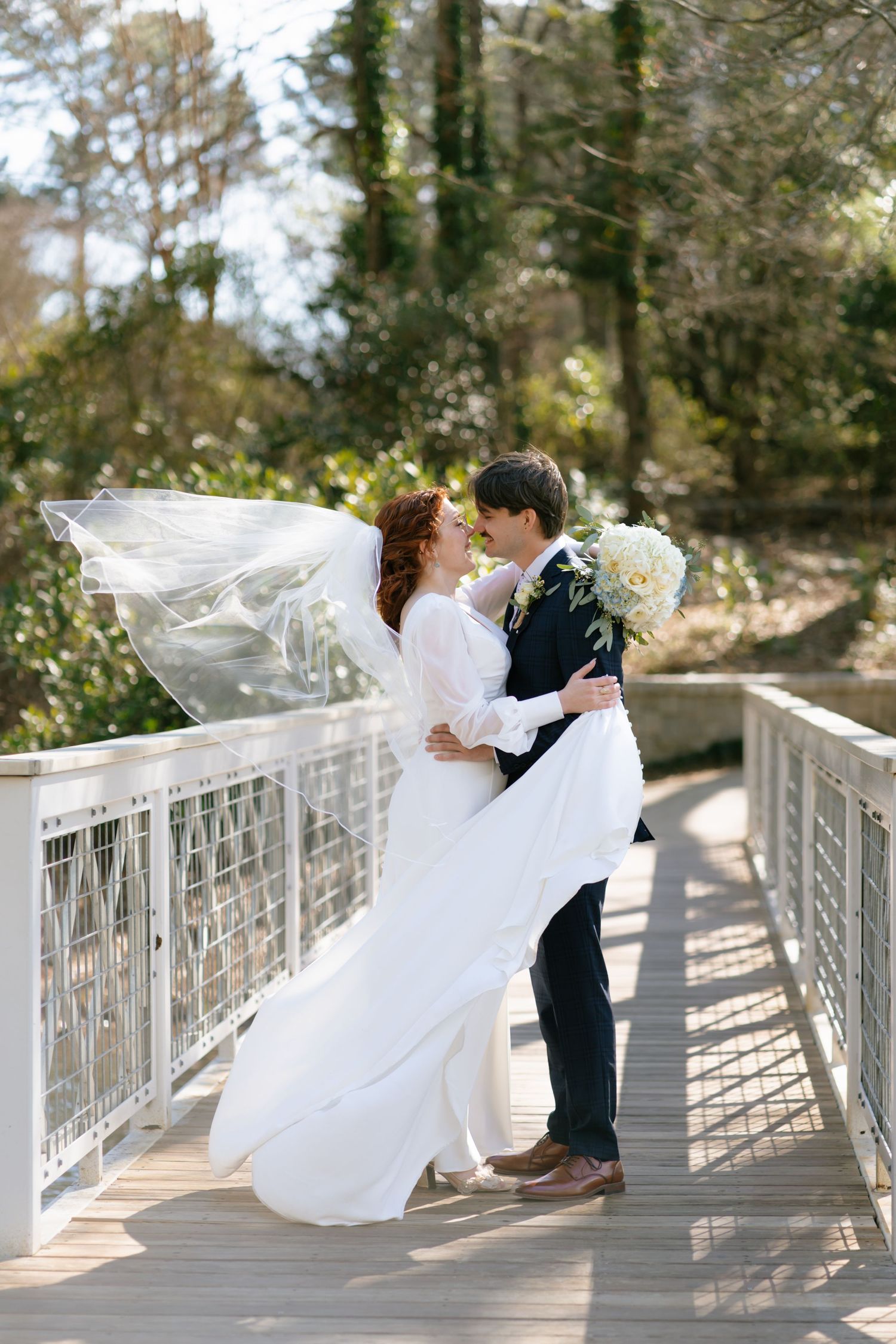 Groom lifts bride in flowing white dress and veil on wooden bridge surrounded by lush trees.
