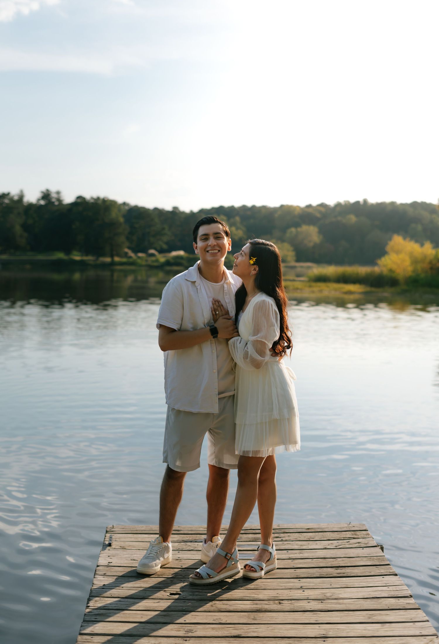 A couple stands on a wooden dock by a calm lake, sharing a romantic moment at golden hour.