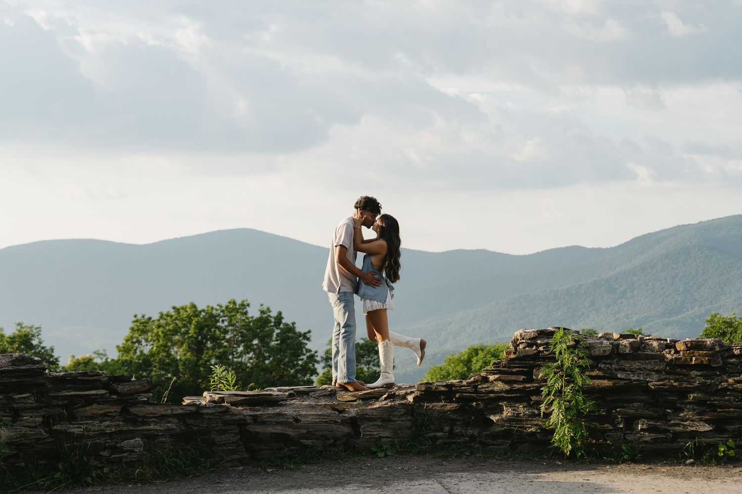 Couple shares a romantic kiss on a stone wall overlook with scenic mountain landscape behind them.