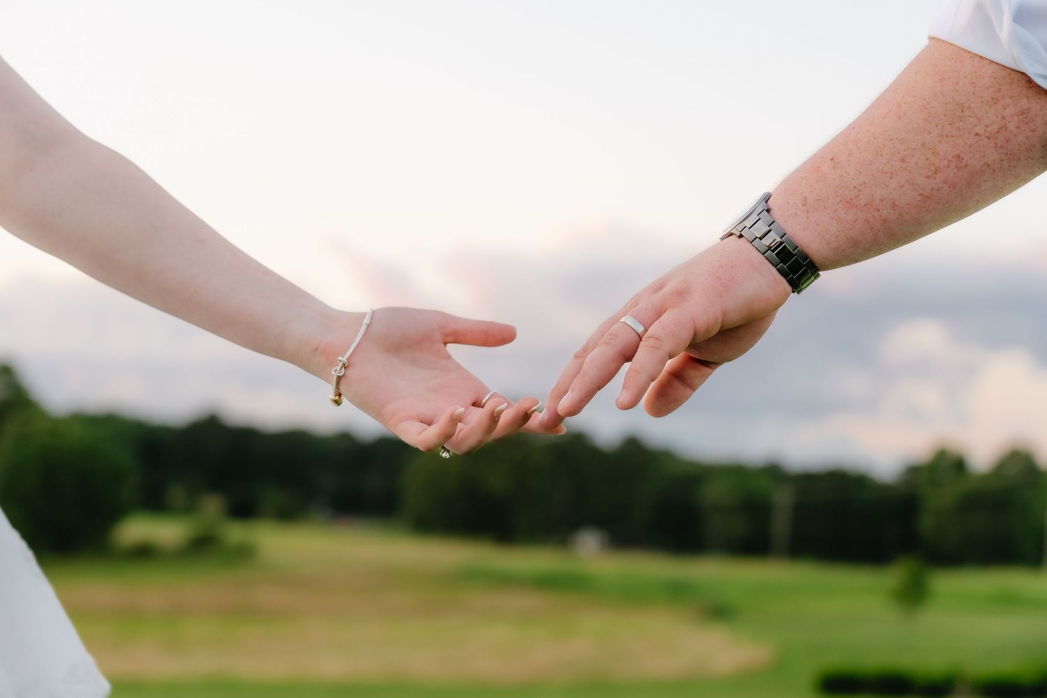 Couple gently reaching for each other's hands outdoors with a green field and cloudy sky in the background.