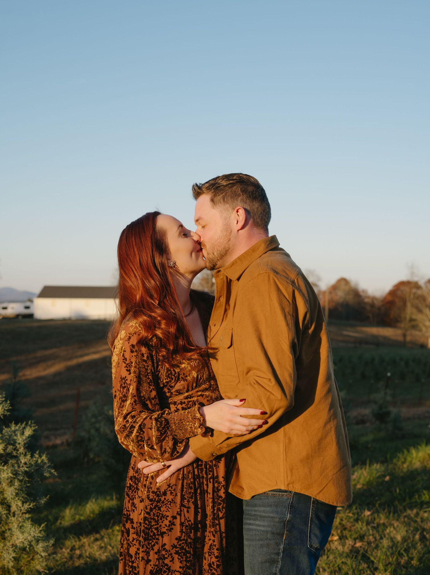 Couple sharing a romantic kiss at golden hour in a rural farm setting, woman in floral dress, man in mustard shirt.