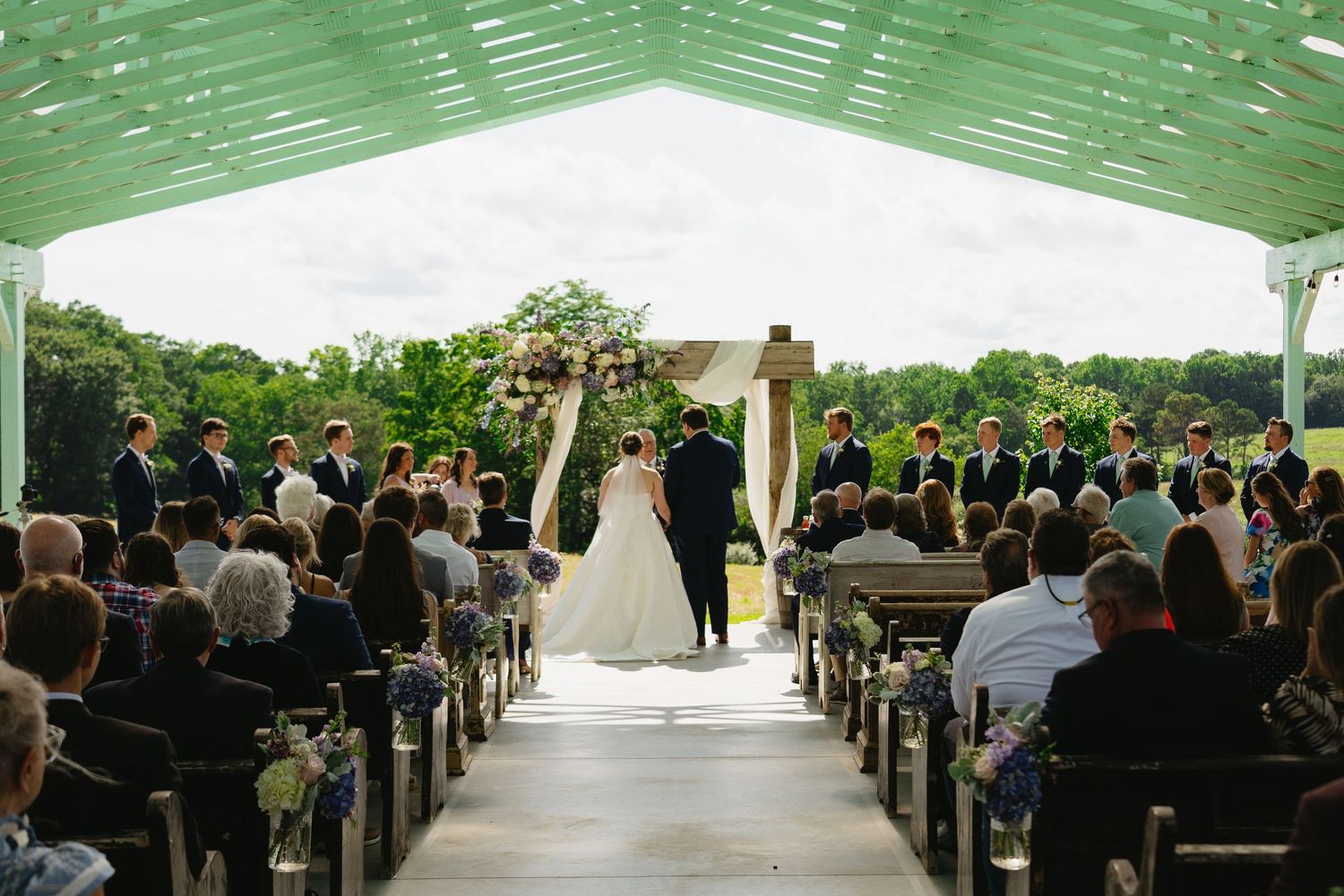 Elegant outdoor wedding ceremony under a white tent with guests seated along an aisle leading to a floral arch altar.