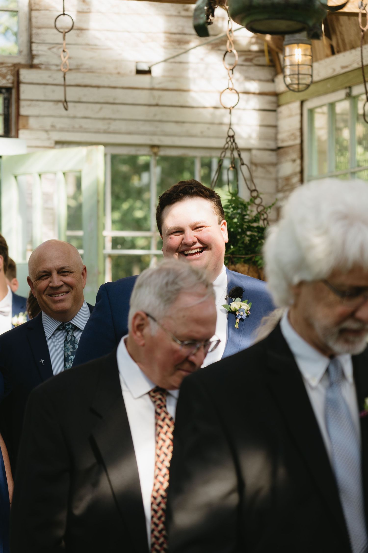Smiling groom in blue suit with boutonniere laughing with wedding guests in rustic venue.