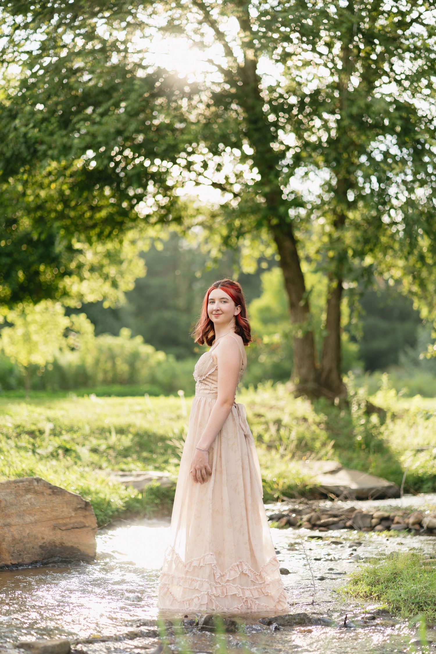 A smiling young woman with red hair wears a flowing beige dress while standing in a sunlit creek.