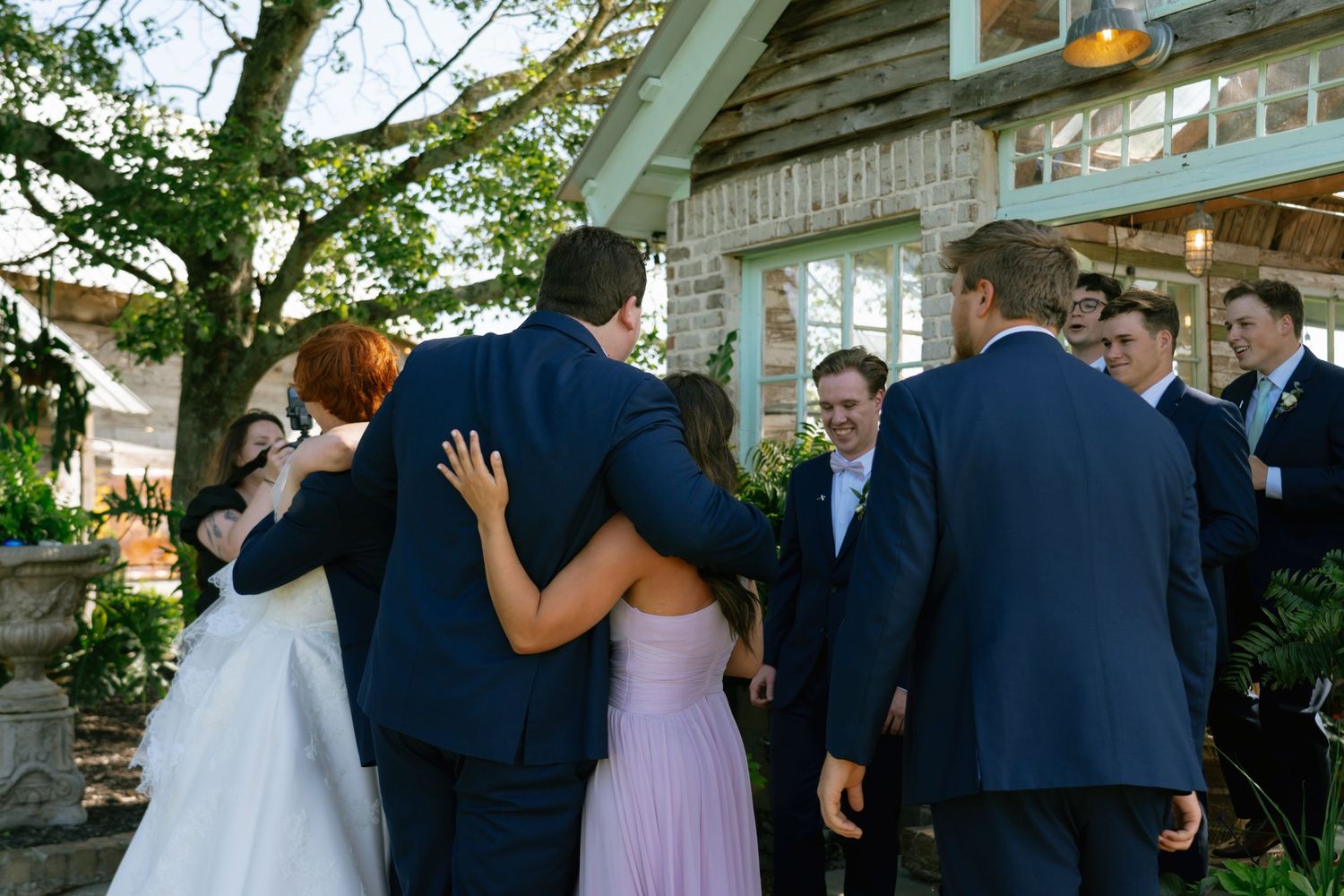 Wedding guests gather and embrace outdoors near a rustic brick venue surrounded by lush greenery.