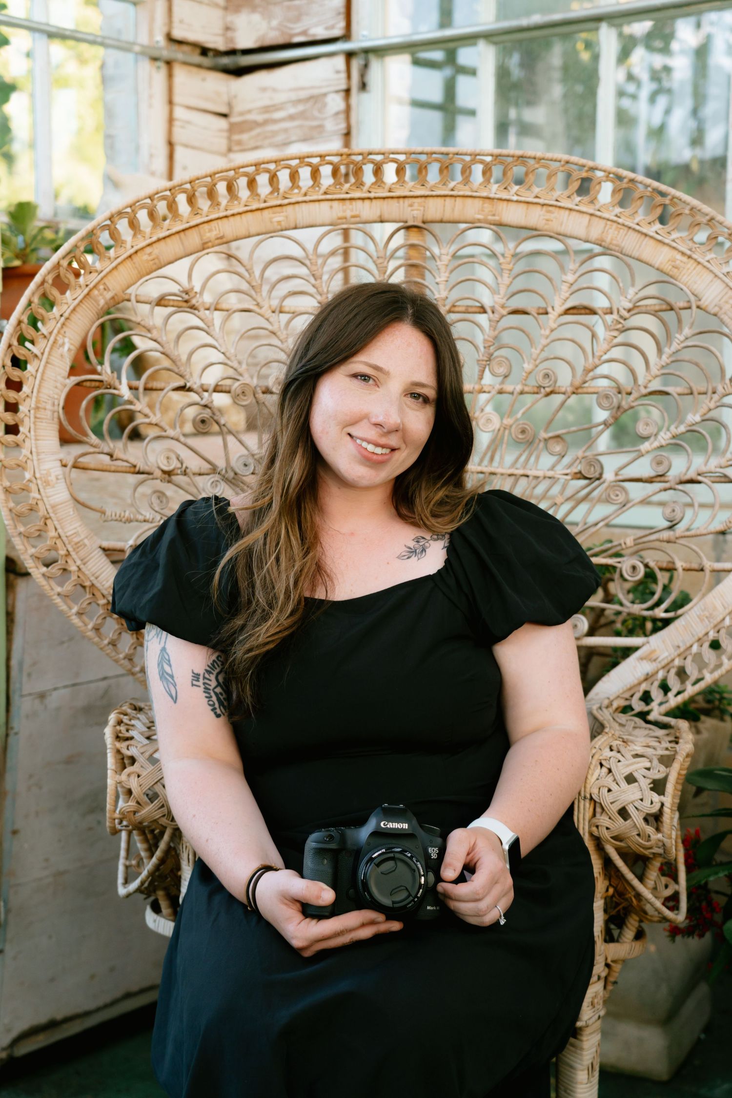 Woman in black dress holding Canon camera seated in wicker peacock chair, smiling outdoors.