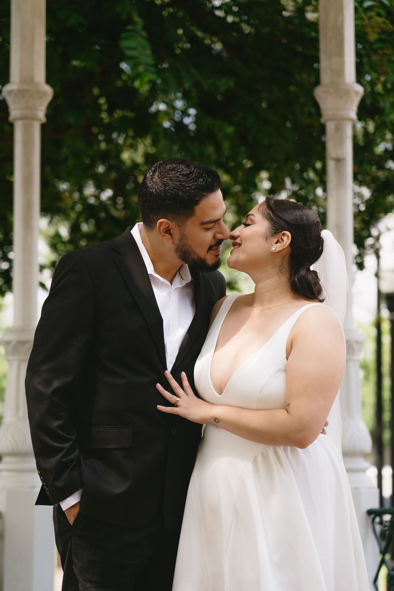 A bride and groom share a romantic nose-to-nose moment under a white gazebo surrounded by greenery.