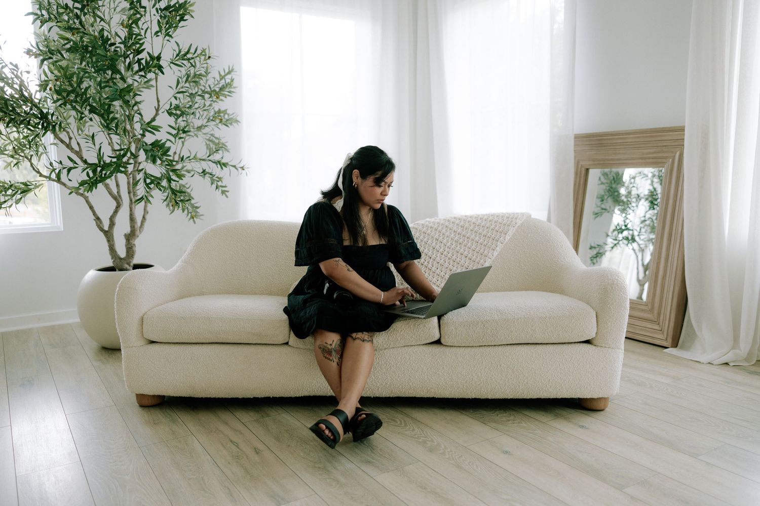 Woman sitting on white sofa using laptop with headphones in bright living room.
