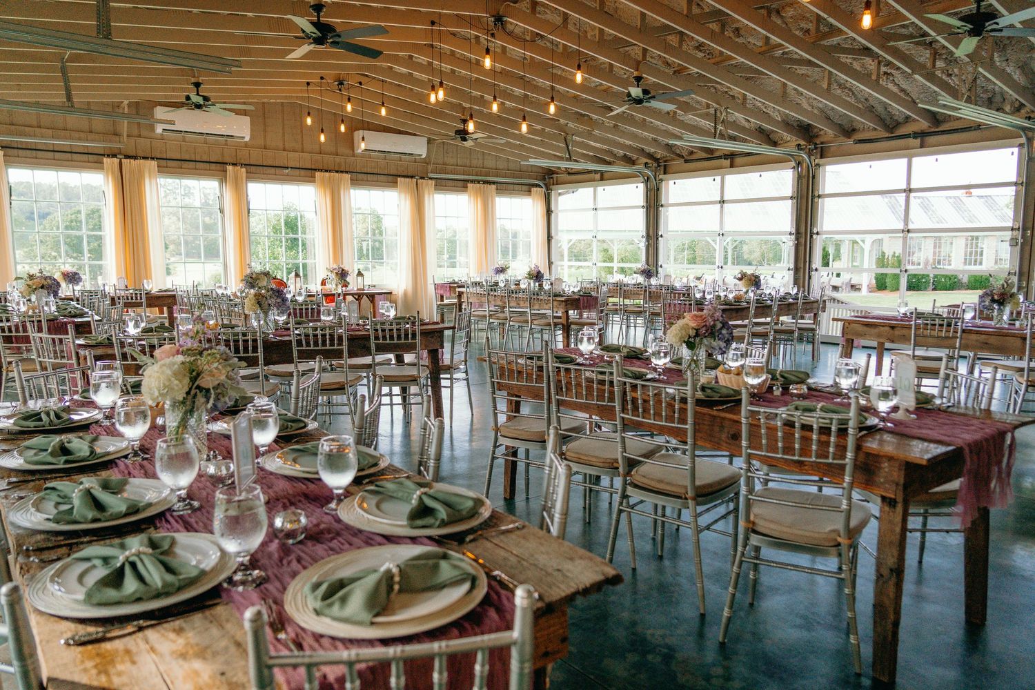 Bright wedding reception hall with round tables set with green napkins, floral centerpieces, and string lights.