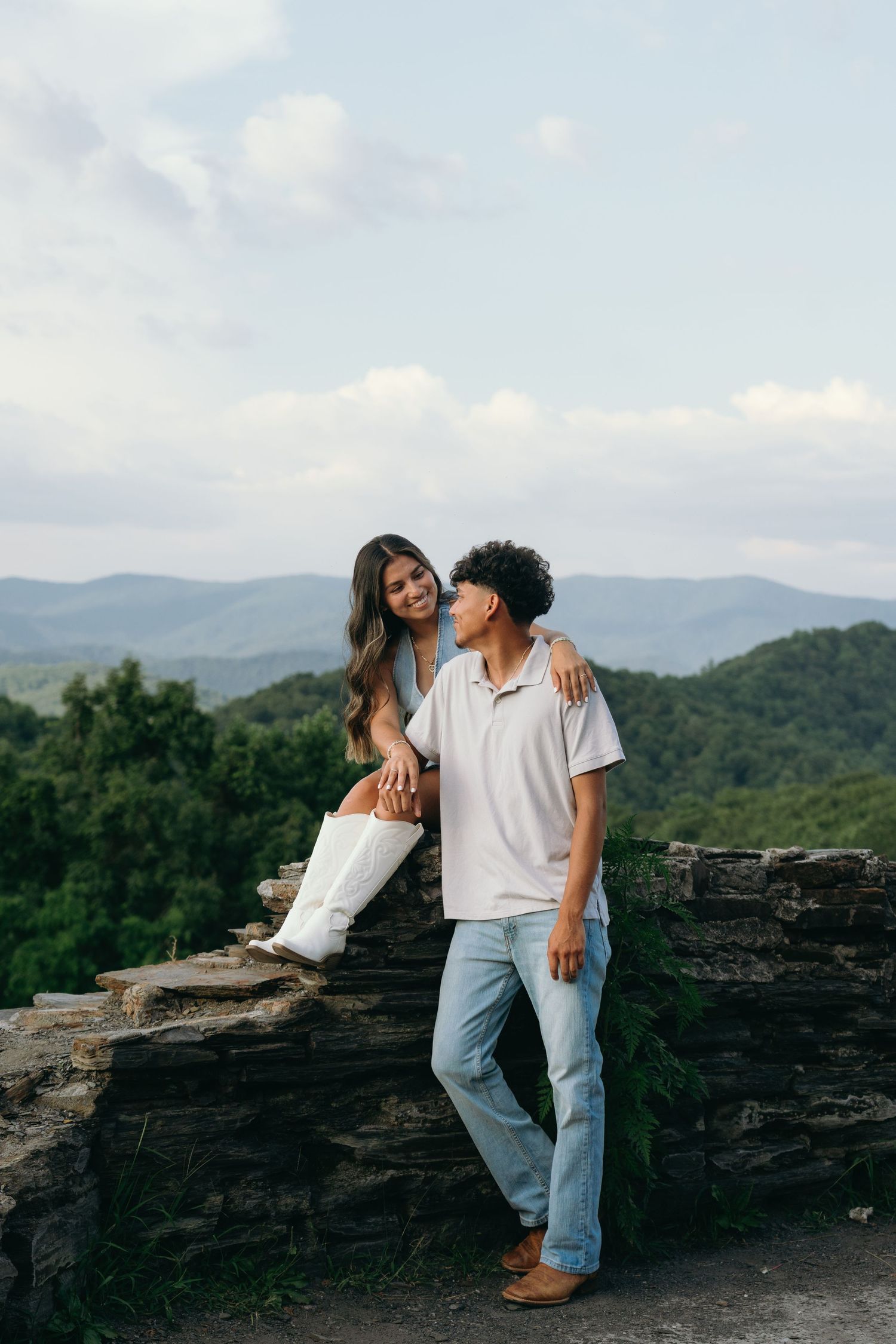 A couple poses together on a stone wall overlook, smiling, with lush green mountains behind them.