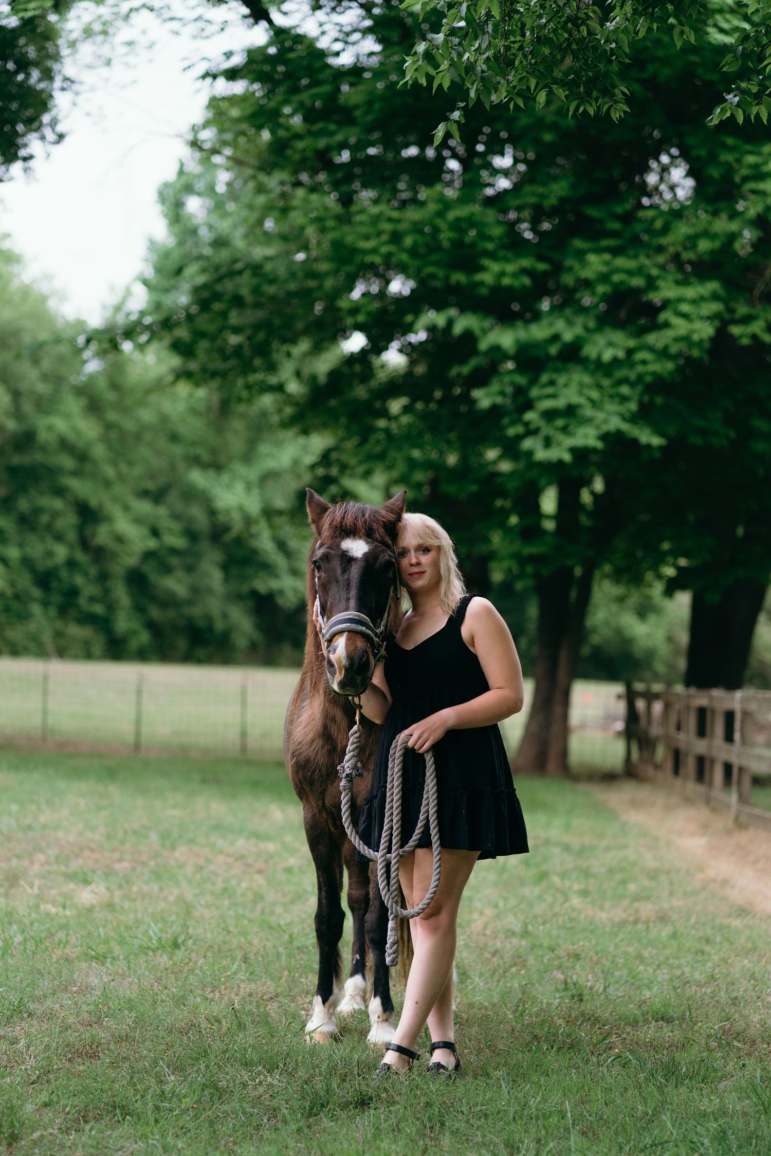 Blonde woman in black dress stands beside dark brown horse with white blaze in lush green tree-lined paddock.