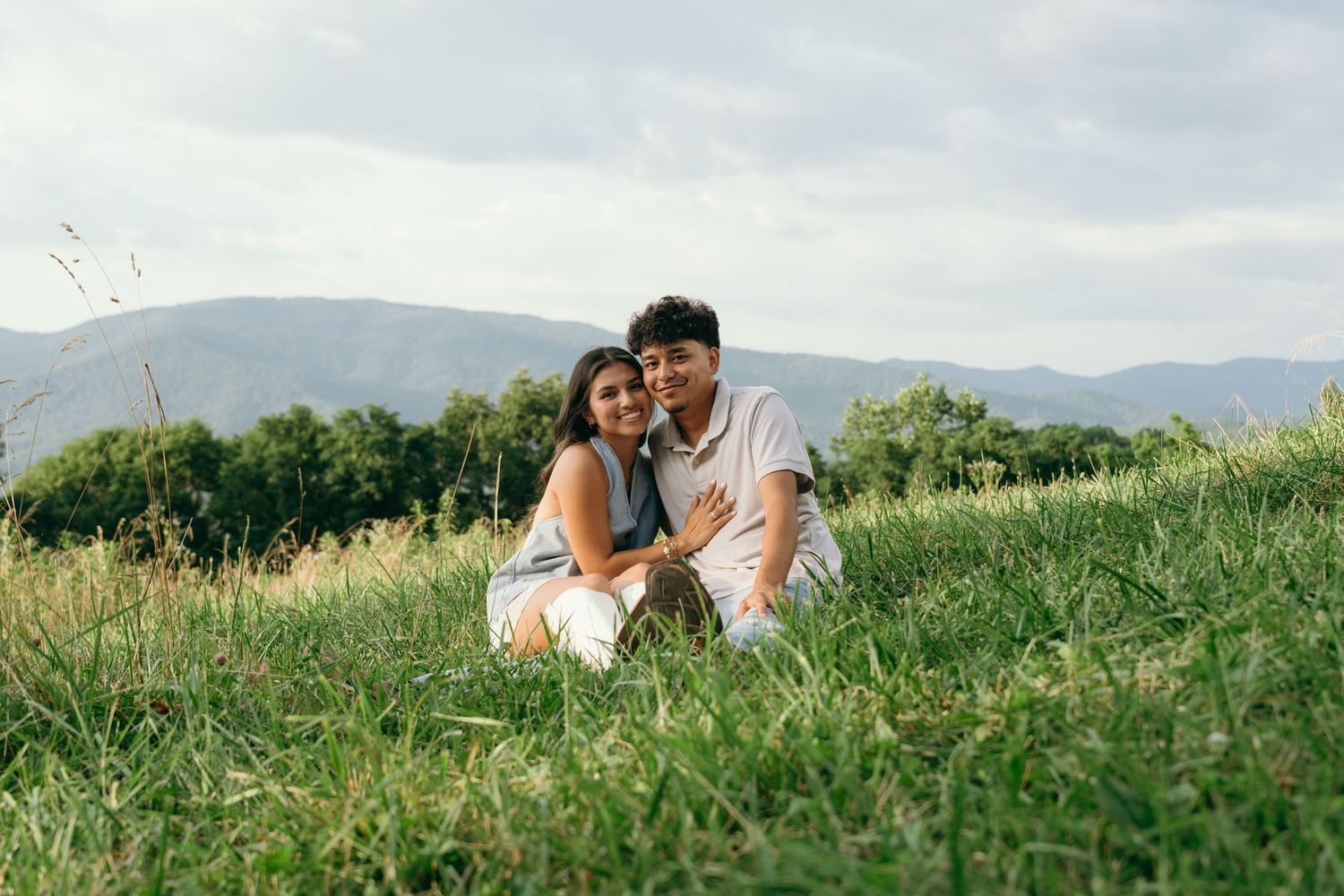 A couple sits together on a grassy hillside with mountain views and green trees in the background.